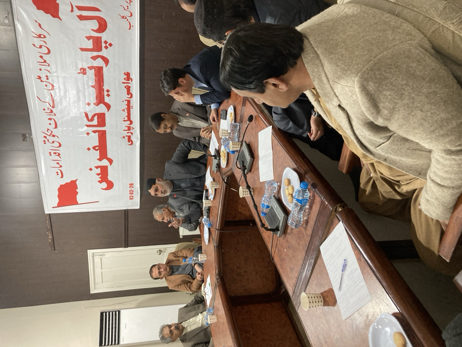 An indoor political meeting is depicted, taking place in a conference room identified by a banner as an "All Parties Conference" at a "Press Club," inferred to be located in Quetta City Tehsil, Pakistan. The banner, dated "03-02-26," also indicates involvement of the "Awami National Party" and addresses "Government measures against government employees."
Approximately ten adult males are visible, seated around a U-shaped, polished dark wooden table. Most individuals are dressed in business-casual attire, predominantly blazers over traditional Pakistani garments. One central male figure with a dark beard and cap is actively speaking, gesturing with his right hand, positioned behind a tabletop microphone. Other attendees are observed listening attentively, with some looking towards the speaker.
The table surfaces contain multiple items: tabletop microphones, plastic bottles of mineral water, white ceramic plates holding biscuits, loose papers, and pens. The room's environment includes dark wooden wall paneling, a white vertical air conditioning unit on the left wall, and a closed white door in the background. The scene suggests a formal discussion or presentation among the participants.