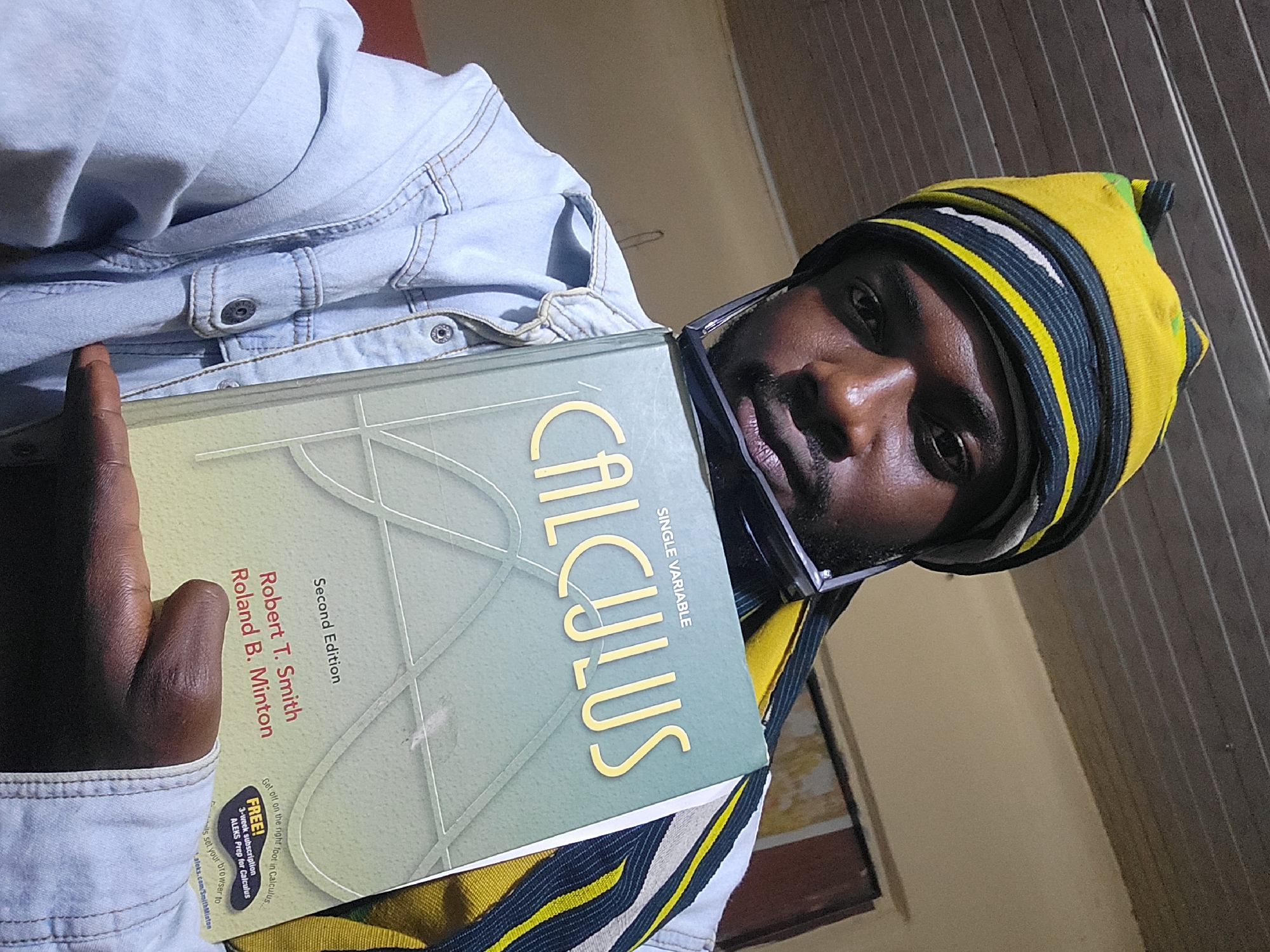 A young person, likely a male, with dark skin, is depicted in a close-up, angled shot, appearing to take a selfie or self-portrait indoors in Jos, Nigeria. He is wearing a light blue denim jacket and a distinctive yellow, black, and white striped beanie or cap. His face, partially visible, looks directly towards the camera with a neutral to slightly serious expression; eyeglasses frames are also visible near his face.
He is holding a "CALCULUS Single Variable" textbook with a light green/teal cover featuring geometric patterns. The book is titled "CALCULUS" in large yellow letters and indicates it is the "Second Edition" by "Robert T. Smith" and "Roland B. Minton." A sticker or printed section on the book offers a "FREE! 3-week subscription ALEKS Prep for Calculus" and directs users to "aleks.com/SmithMinton" to "Get off on the right foot in Calculus..."
The background consists of plain, light-colored walls, with one section appearing to have vertical paneling or blinds. The overall lighting suggests an indoor setting, possibly with natural light. The image is rotated counter-clockwise, giving it a dynamic, informal feel.