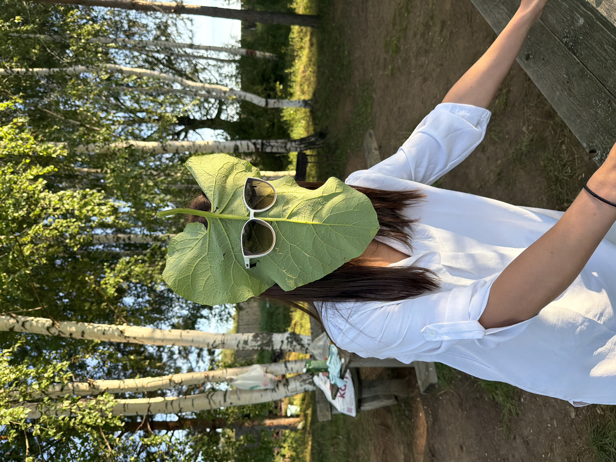 A person wearing a white shirt and sunglasses is holding a large green leaf in front of their face. The person is outdoors, in what appears to be a park or wooded area. The background consists of several birch trees with green foliage. A picnic table with some items on it is visible to the left, and a wooden bench is in the foreground. The lighting suggests it is daytime, with sunlight filtering through the trees. The location is identified as Dubrova, Russia.