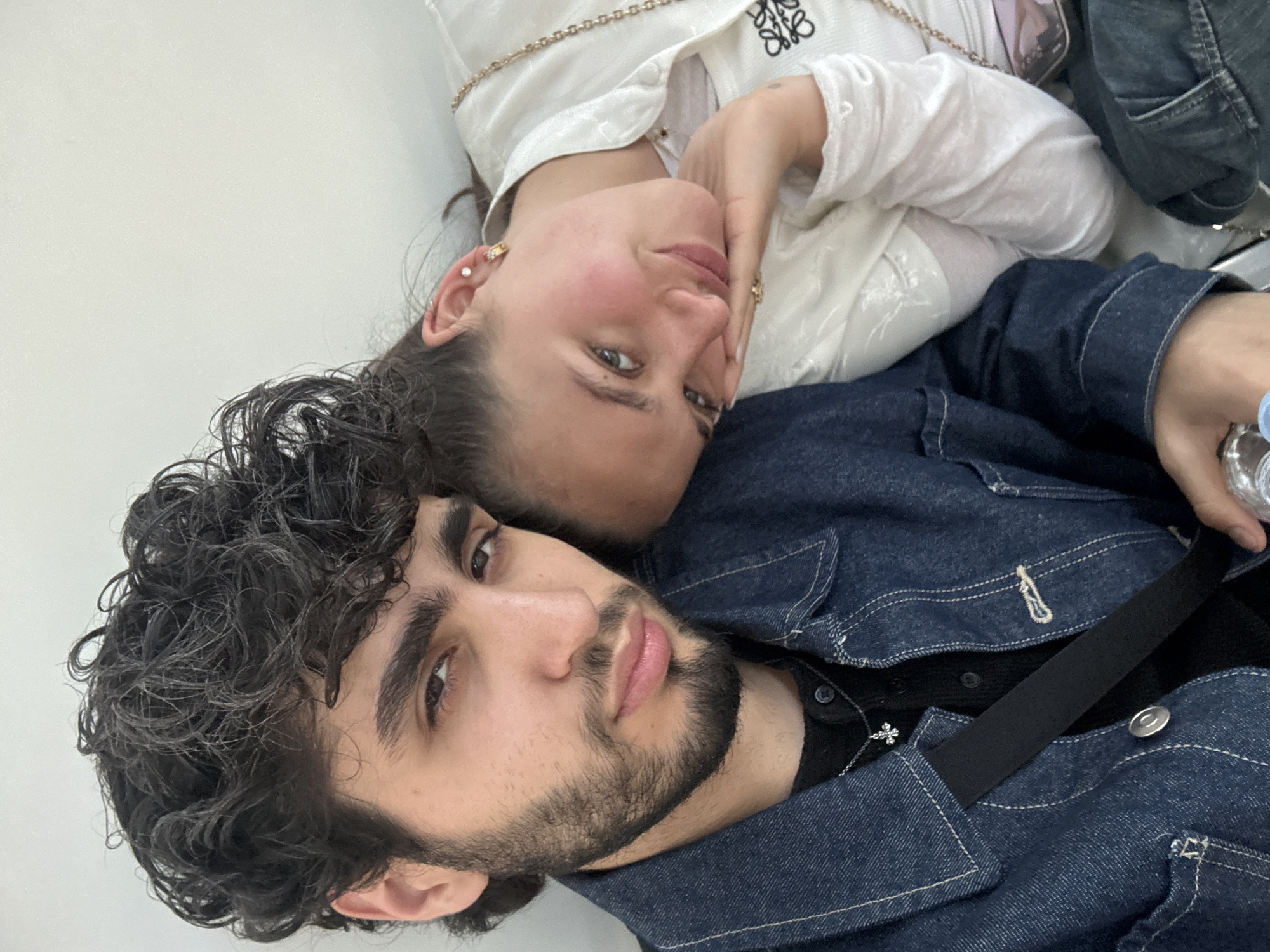 A medium-angle, unedited photograph depicts a couple posing indoors against a solid white backdrop.
The man has curly dark hair and a beard and is wearing a dark denim jacket over a black shirt. He appears to be lying down, with a black strap visible diagonally across his torso.
The woman lies on top of the man, with her head resting close to his. She has brown hair pulled back and is wearing a white blouse. Her right hand is touching her face.
There is no discernible evidence to suggest this image was taken in Yerevan, Armenia. No specific landmark or detail is present that could pinpoint the location.
