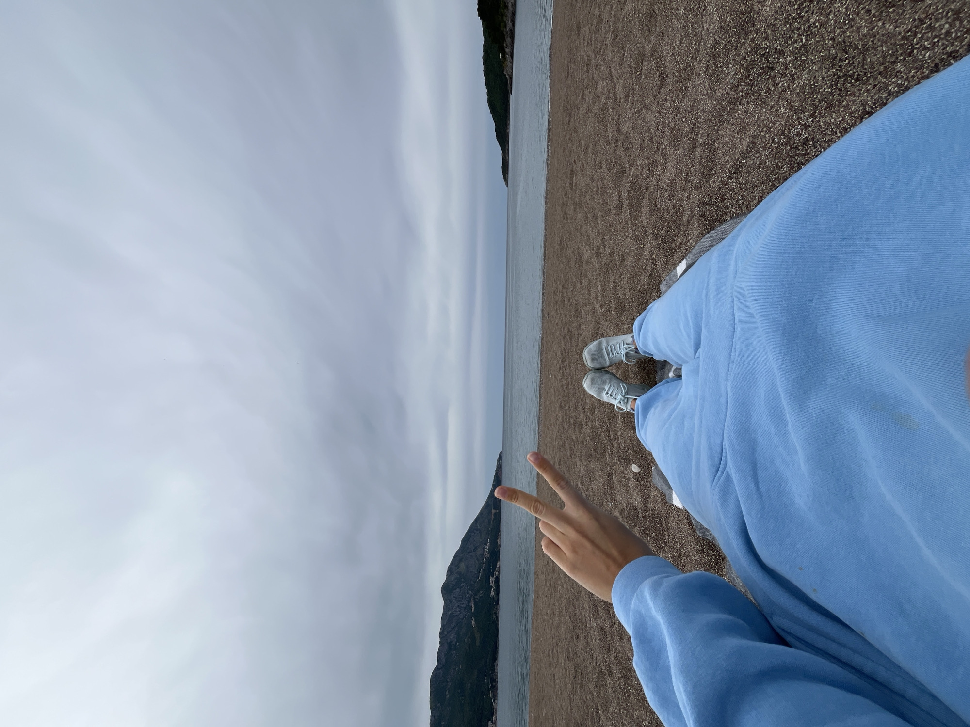 The image, taken from a high angle, features a person dressed in a light blue tracksuit and grey sneakers, sitting on a beach in Boreti, Montenegro. The sea is in the middle ground of the image, and in the background, there are mountains and a cloudy sky. The person in the image is making a peace sign with their hand. The beach appears to be made of small pebbles.