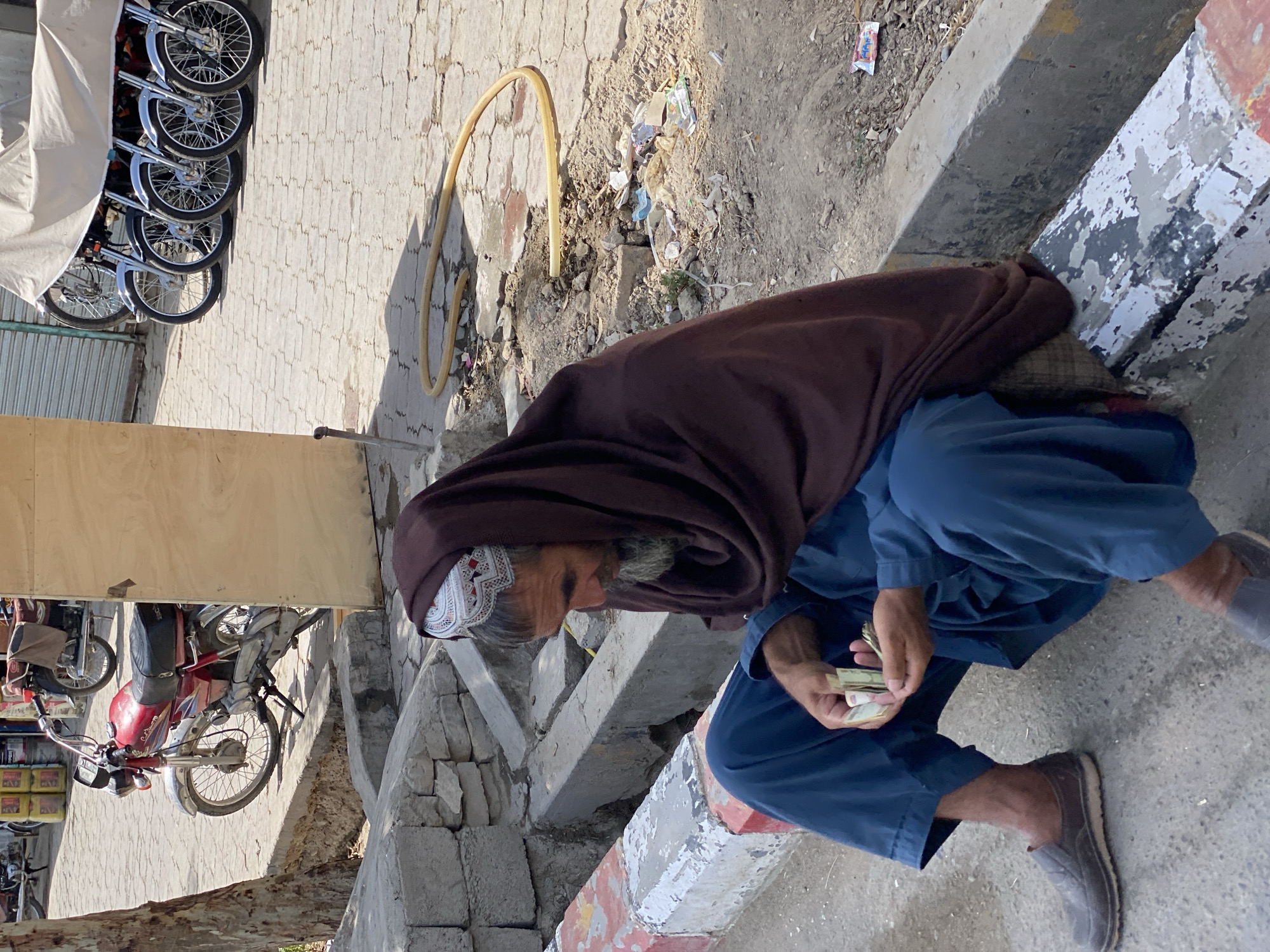 An elderly man with a gray beard and a white and red embroidered cap sits on the side of a street in Kandahar, Afghanistan. He is wearing a brown shawl draped over his head and shoulders and a loose-fitting blue garment. He is holding a small wad of cash in his hands and appears to be counting or sorting it. The street is paved with interlocking stones and there is a yellow hose lying on the ground behind him. Several motorcycles are parked in the background, some covered with a tan tarp. The scene appears to be in a commercial area with some debris and scattered trash on the ground. The lighting suggests it is daytime with the sun casting shadows.