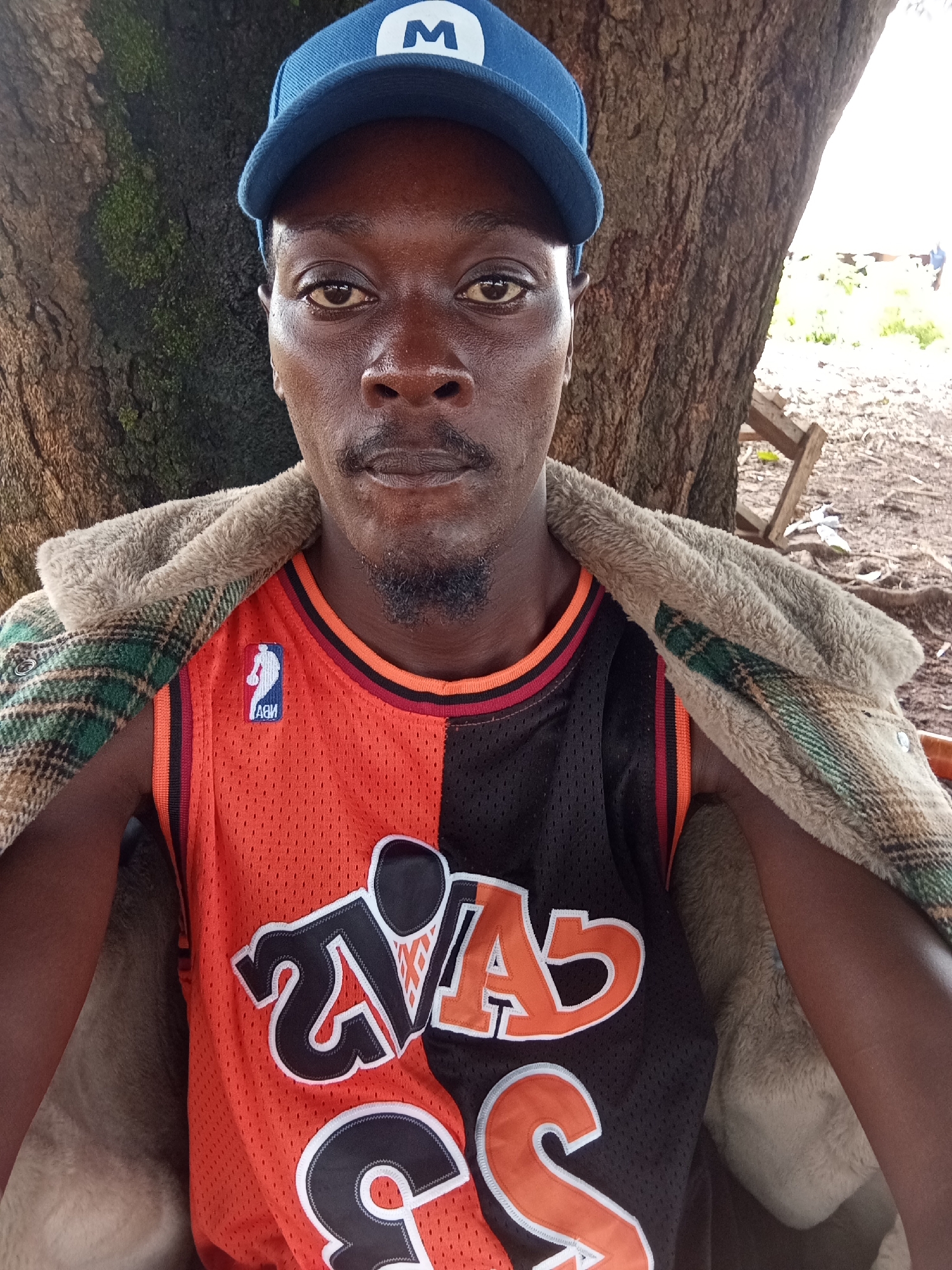 A medium shot captures a Black man wearing a blue baseball cap with a white 'M' logo, a tan and green plaid jacket draped over his shoulders, and an orange and black basketball jersey with the NBA logo. The man has dark skin, a goatee, and is looking directly at the camera. He is seated in front of a large tree trunk with rough bark, its branches and leaves visible in the background. The ground is covered in dirt and some scattered debris. The overall setting appears to be outdoors, possibly in a rural or semi-rural area, with a wooden structure visible in the distance. The lighting suggests it is daytime. The man is the primary subject, and the image is a selfie or a photo taken by someone close to him. The background provides context of an outdoor environment.
