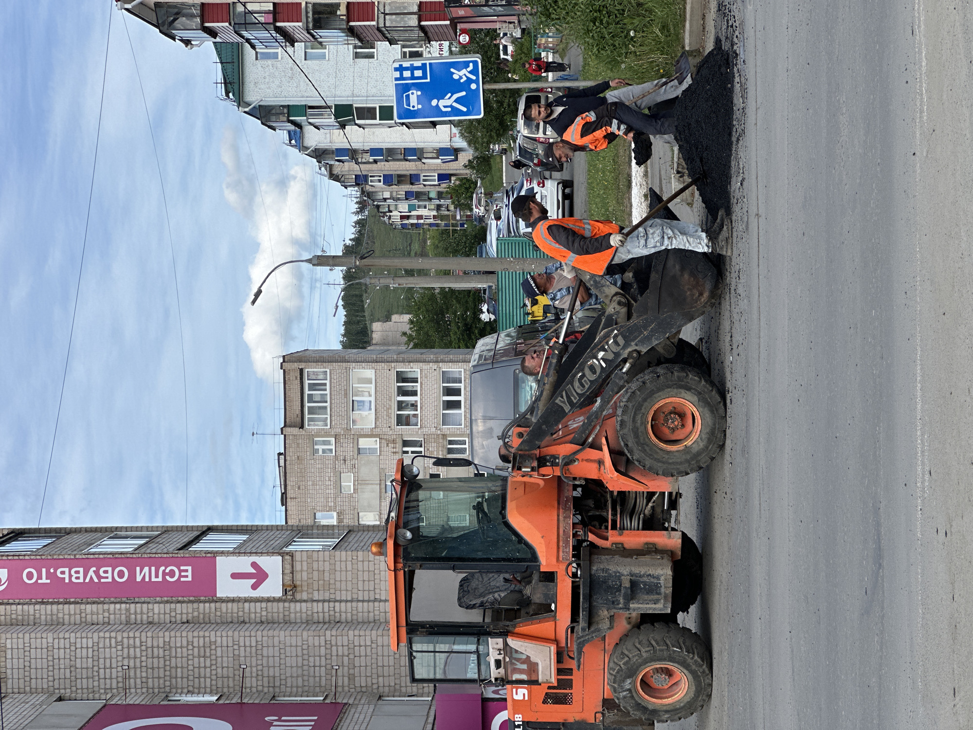 The image captures a street maintenance scene in an urban area. In the foreground, an orange "YIGONG" branded compact wheel loader, with a visible "SOYUZSPETSOTECH" sticker, is parked on the asphalt road. A team of road workers, all wearing orange vests, are actively involved in the asphalt repair. One worker is shoveling asphalt from the loader's bucket onto the road surface, while another is spreading it. The scene is set against a backdrop of city buildings, including multi-story apartment blocks with diverse architectural elements such as balconies, windows, and varying exterior designs. A street sign indicating a pedestrian crossing is also visible. The sky is partly cloudy.