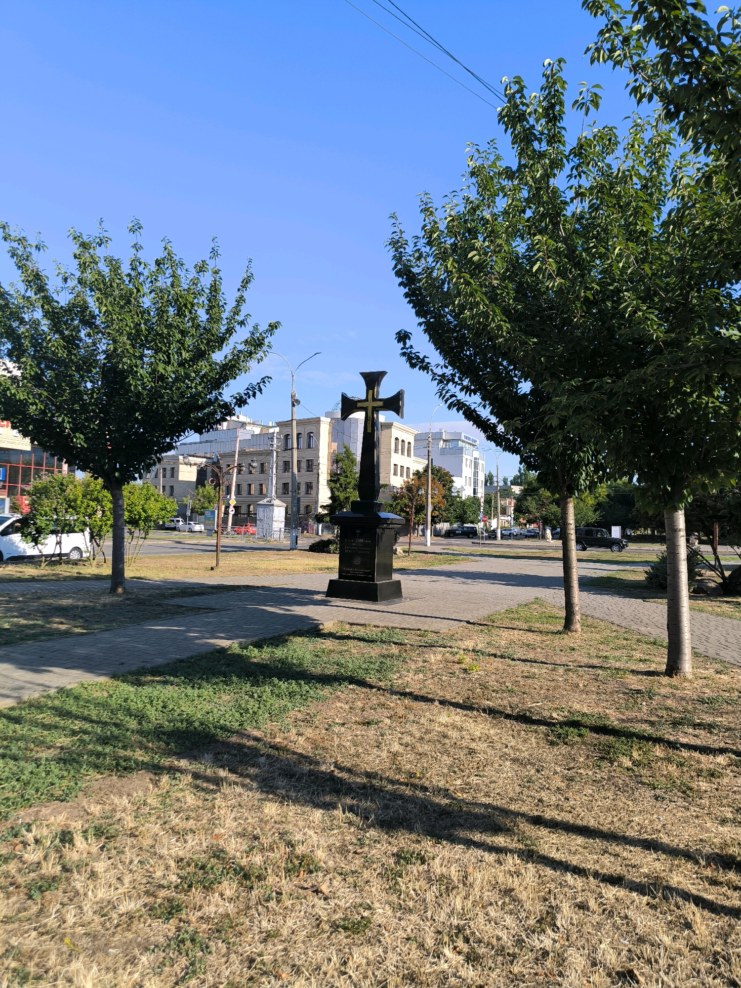 The image depicts a monument with a cross on a pedestal, situated in a public space in Mykolaiv, Ukraine. The monument is located on a paved walkway, with grass and dry vegetation in the foreground. Two trees flank the monument, with their shadows cast across the grass. In the background, there are buildings, a street with cars, and utility lines overhead. The sky is clear and blue, indicating a sunny day. The monument appears to be made of dark stone with a golden cross. The inscription on the pedestal is partially visible. The overall scene suggests a well-maintained urban environment.