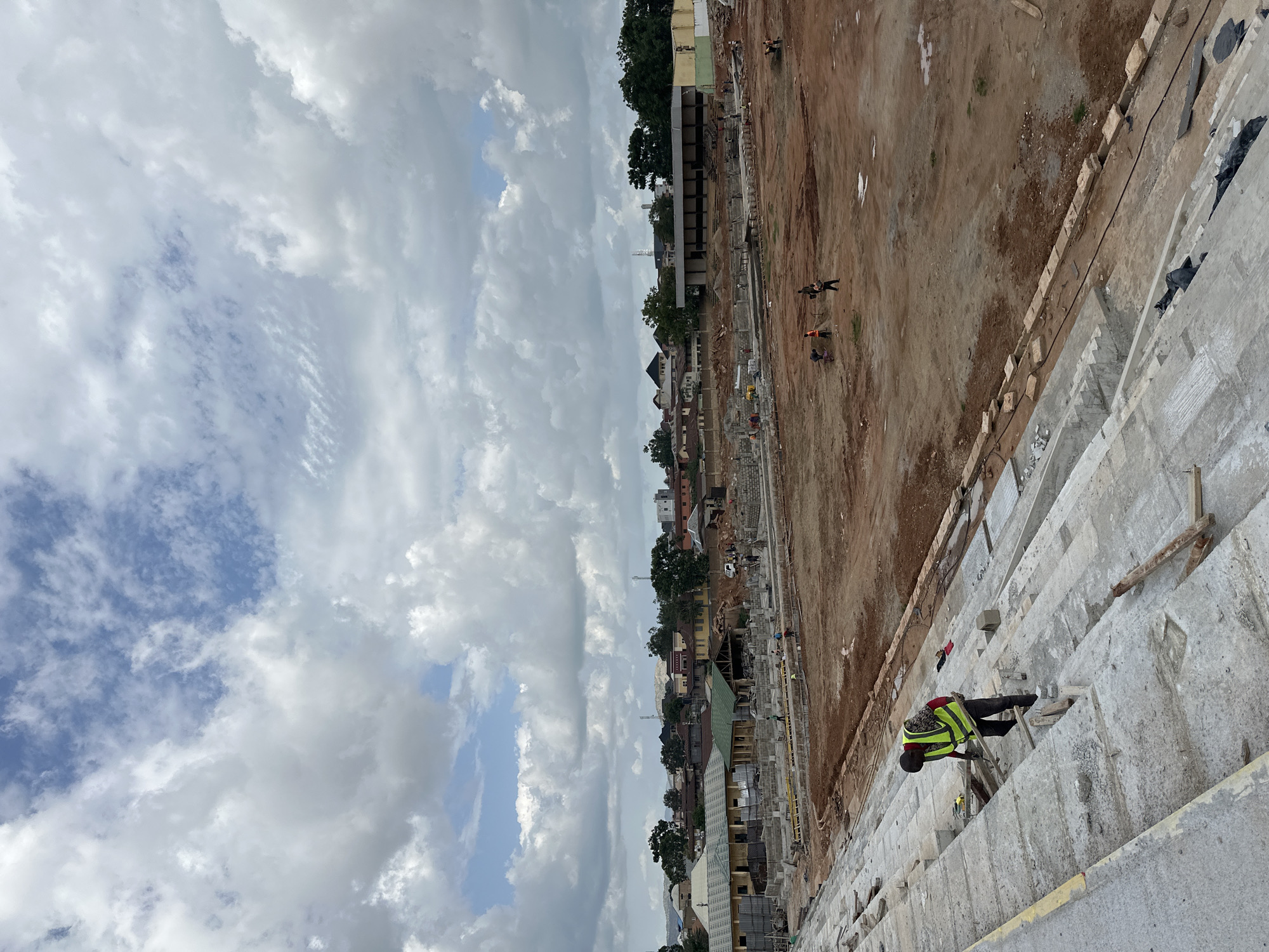 The image shows a construction site with several people working. The scene is outdoors, under a cloudy sky, suggesting it's daytime. The ground is mostly dirt and some concrete structures are visible. In the foreground, a person wearing a high-visibility vest and dark pants is standing on wooden scaffolding, appearing to be engaged in some form of construction work. Further back, other individuals are scattered across the site, some walking and others seemingly working.
To the left of the construction area, there is a dense collection of buildings, appearing to be a residential or mixed-use area, with varying roof colors and architectural styles. Trees are interspersed among the buildings. The sky is filled with a mix of white and gray clouds, with patches of blue sky visible, indicating a partly cloudy or overcast day. The overall impression is a busy construction project in an urban or semi-urban environment. No visible text is present in the image.