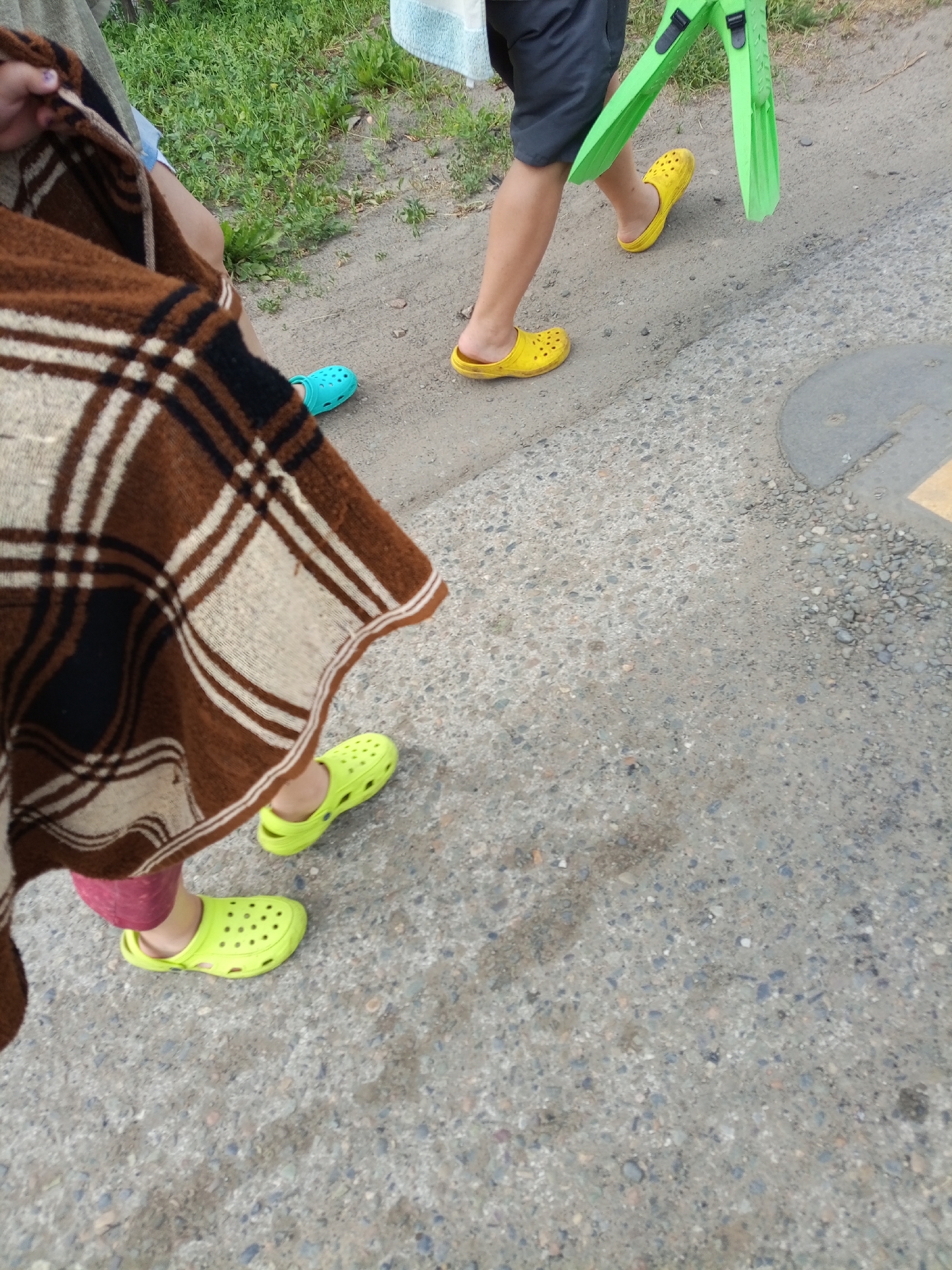 In Pankrushikha, Russia, two individuals are walking outdoors on a grey, pebbly path. The person in the foreground, partially obscured by a brown and white plaid blanket, wears lime green Crocs and pink leggings. Ahead of them, another person, wearing black shorts, walks in bright yellow Crocs and carries a pair of green swim fins. A third individual, whose feet are not visible, is also present and wears bright blue Crocs. Green grass borders the path to the left.