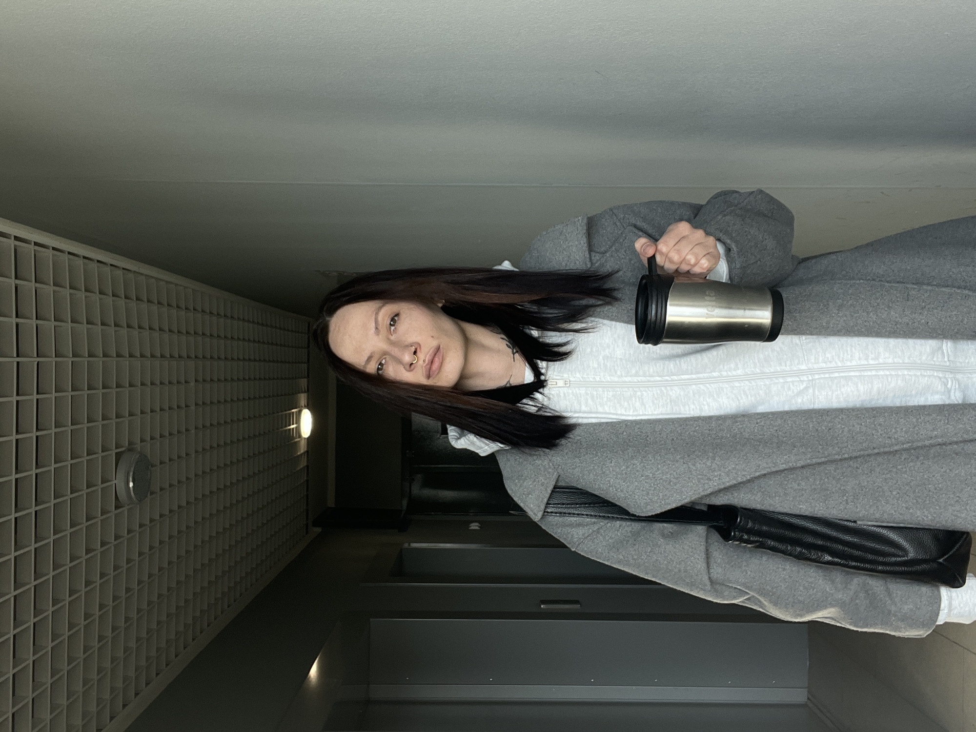 A young woman stands indoors, holding a silver mug. She is wearing a grey coat over a white zippered hoodie. A black leather purse hangs from her shoulder. The location appears to be a modern building with a grid-like wall design and recessed lighting. Based on the style of the architecture, this image might be from a commercial or residential building in Izhevsk, Russia. However, without additional information or visual cues characteristic of Izhevsk, definitive location identification is impossible.
