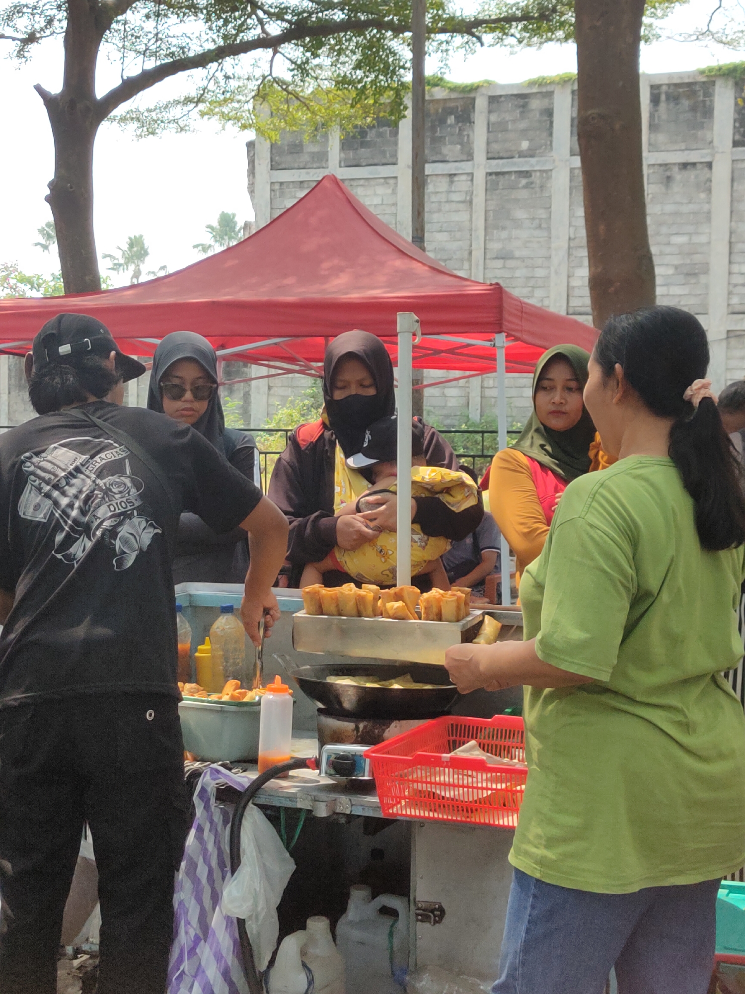 The image depicts an outdoor scene in Ngasem, Indonesia, likely a street food vendor stall. In the foreground, a person wearing a black t-shirt with a graphic print and a black baseball cap is seen from behind, interacting with the food stall. To their left, two women are visible. One, wearing a black hijab and sunglasses, looks towards the stall. Another woman, wearing a black hijab and a black face covering, is holding a yellow object, possibly food. A third woman, wearing a green hijab and a pink and yellow top, is standing to the right, looking at the person in the black t-shirt.
The food stall itself features a red canopy tent. On the stall, there are various items, including a metal tray with what appears to be fried food items, a green plastic container, bottles of condiments (one yellow, one with an orange cap), and a red plastic basket containing more food items wrapped in brown paper. A wok or frying pan is visible on a portable stove behind the food tray. In the background, a white brick wall and trees can be seen. The overall environment suggests a bright, sunny day.