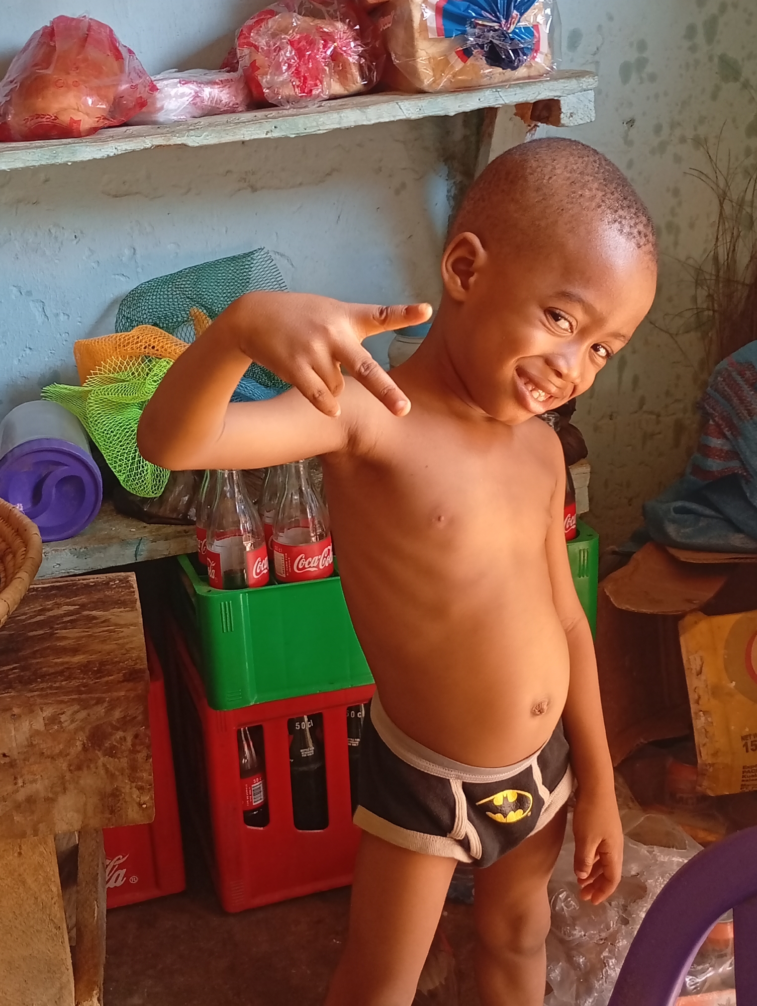 A young, shirtless boy in Yelwa, Nigeria, smiles and makes a "peace" sign with his left hand. He wears black briefs with a yellow Batman logo. Behind him is a shelf holding loaves of bread in plastic bags, a stack of green and red Coca-Cola crates holding glass soda bottles, and a purple container. The room has pale blue walls.