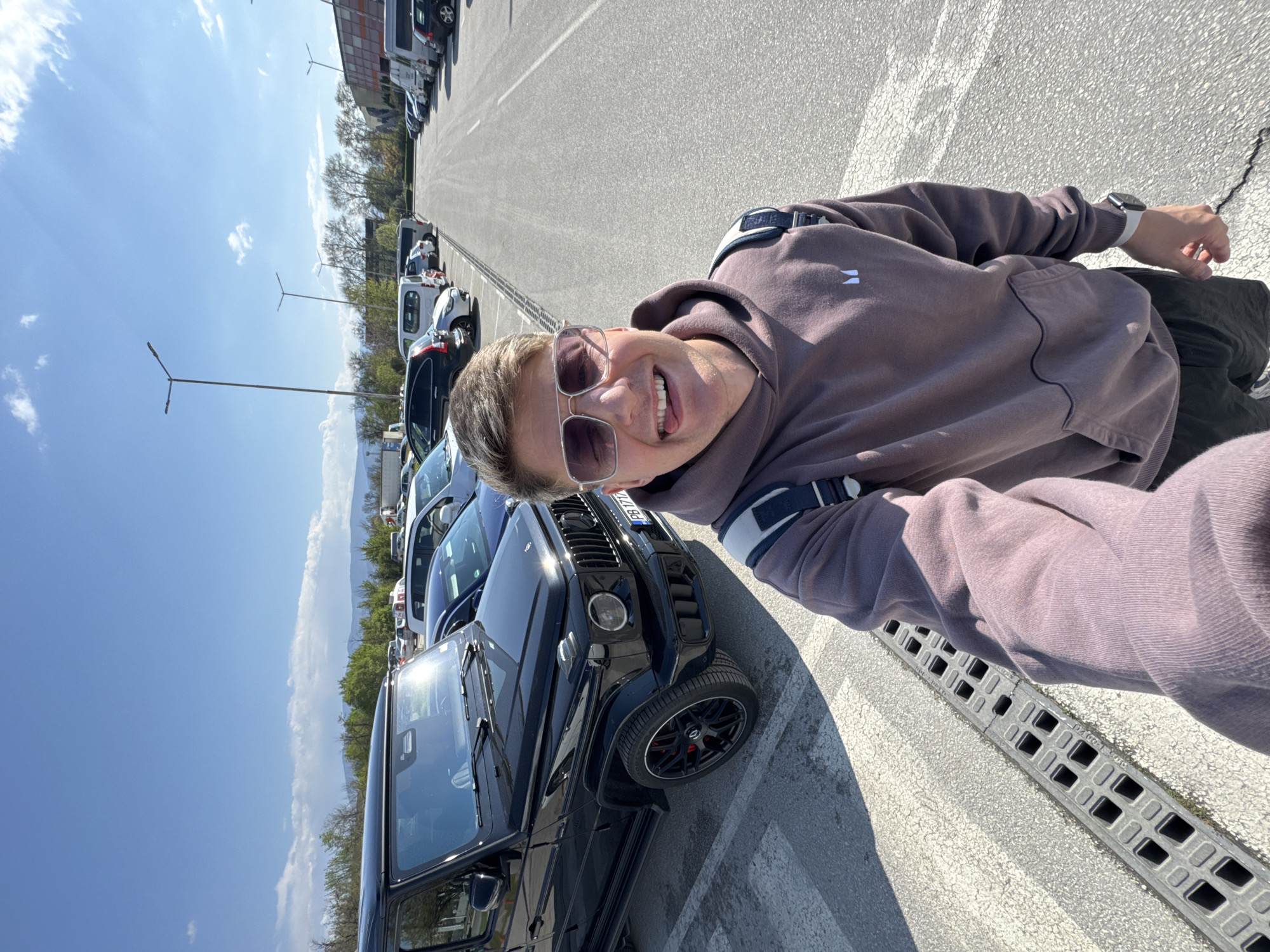 A man is taking a selfie in a parking lot in Plovdiv, Bulgaria. He is wearing sunglasses and a brown hoodie. Behind him is a black Mercedes-Benz G-Class SUV. Other vehicles are parked nearby. The sky is blue with some clouds.