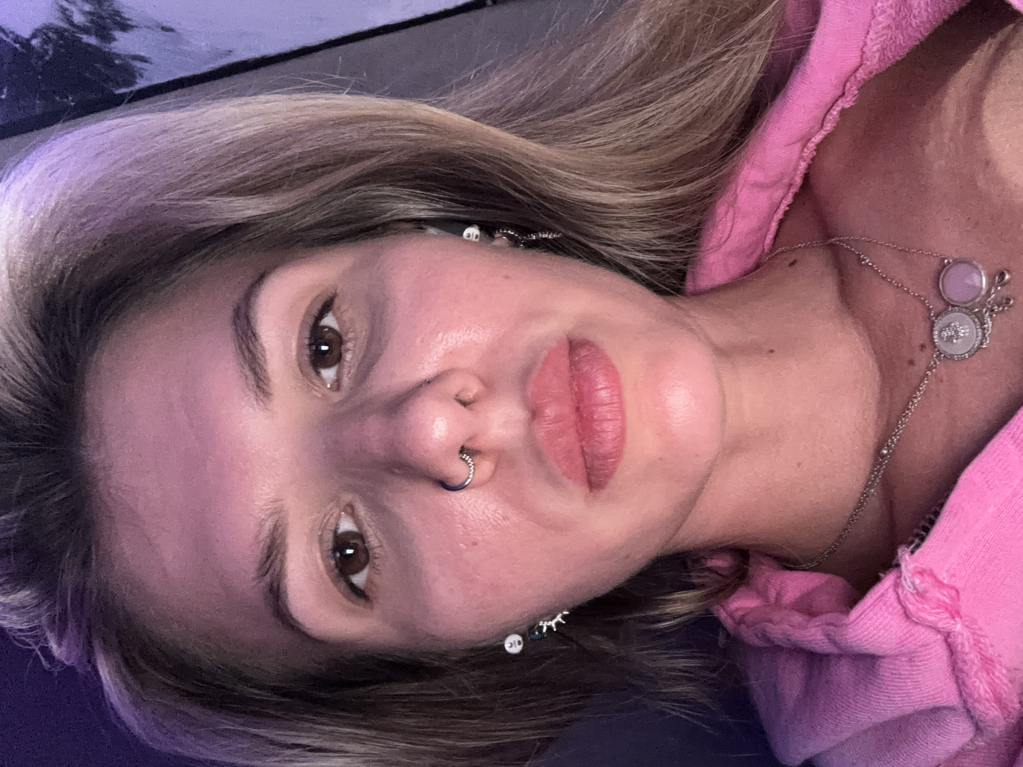 A close-up, head-on, eye-level selfie of a young, Caucasian woman with blonde hair pulled back into a bun, is taken in an indoor setting with purple and blue lighting. The woman has brown eyes, a nose ring, and is wearing a pink hooded jacket and a silver necklace with two pendants. Her skin appears flushed, and she has makeup on her lips. The background is dark and out of focus.