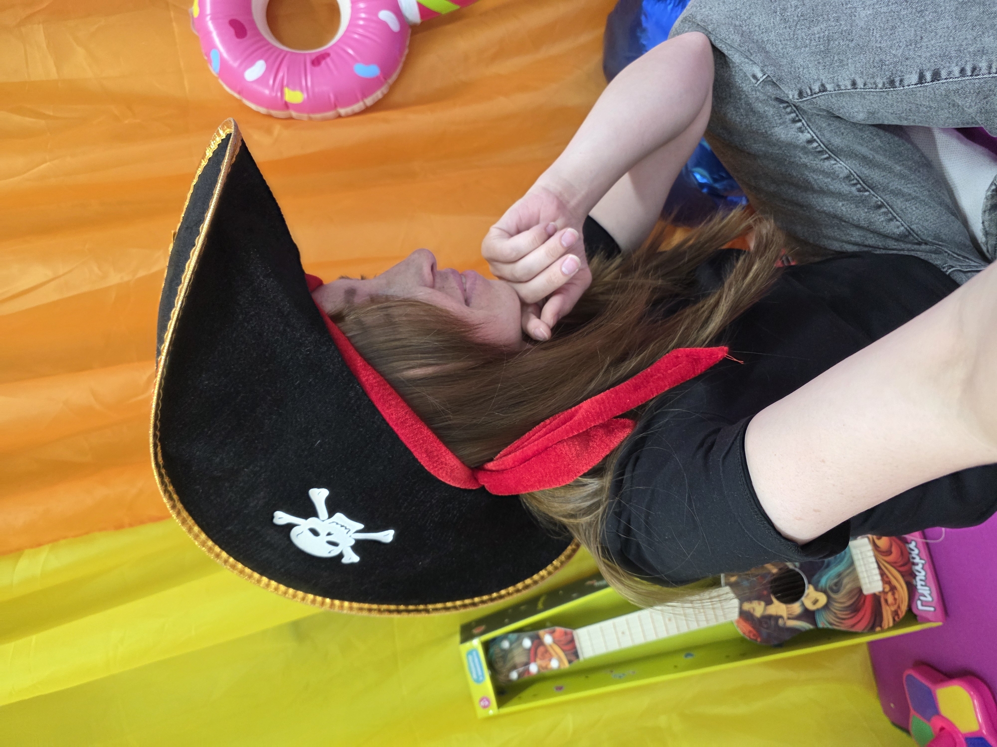This image captures a person in a pirate hat lying down in a brightly colored indoor setting, likely in Chaikovsky, Russia. The individual has long brown hair and is wearing a black t-shirt and grey jeans. A prominent black pirate hat with a red ribbon trim and a white skull and crossbones emblem sits on their head. Their head is turned to the left, with one hand resting near their face, suggesting a relaxed or contemplative posture.
The background is a vibrant, soft surface, transitioning from orange in the upper half to yellow below, resembling a play mat or cushioned floor. Various colorful objects are present: a pink inflatable donut ring with multicolored sprinkles is visible in the upper right. In the lower right, a bright green box holds a toy guitar with a detailed, colorful pattern; the word "Гитара" (Russian for "Guitar") is visible on its packaging. A blue inflatable object is partially seen behind the person's upper body, and a pink toy is also slightly visible in the lower right corner. The scene evokes a playful and casual atmosphere, possibly during a themed event or a relaxed moment in a recreational space.