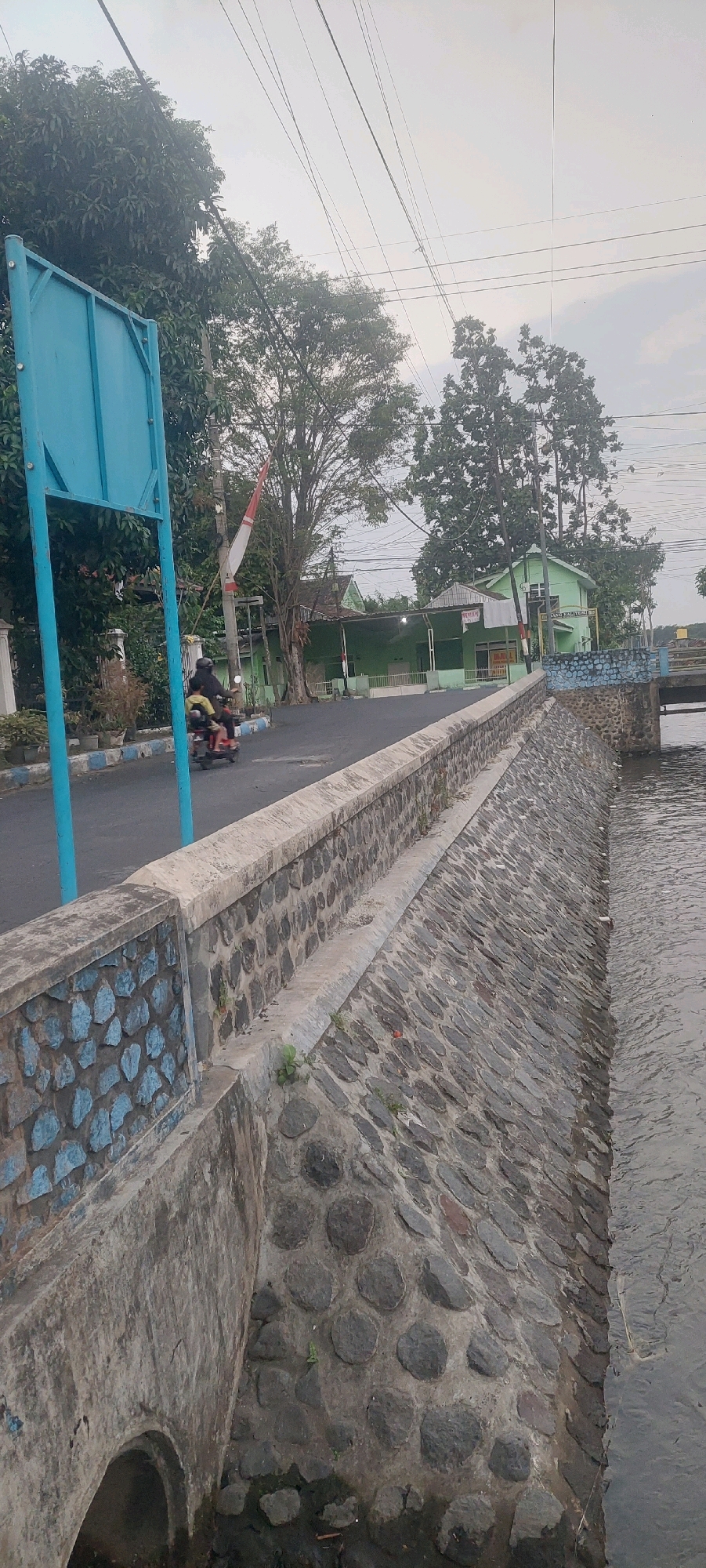 The image depicts a view of a riverbank in Lumajang, Indonesia. A stone embankment lines the right side of the frame, with a river flowing to its right. On the left side, a road is visible, with a motorcycle carrying two people riding on it. A blue metal signpost stands on the left edge of the road. Beyond the road, there are trees and green buildings, with a red and white flag flying from a pole. The sky is overcast. The scene appears to be an urban or semi-urban setting along a waterway.