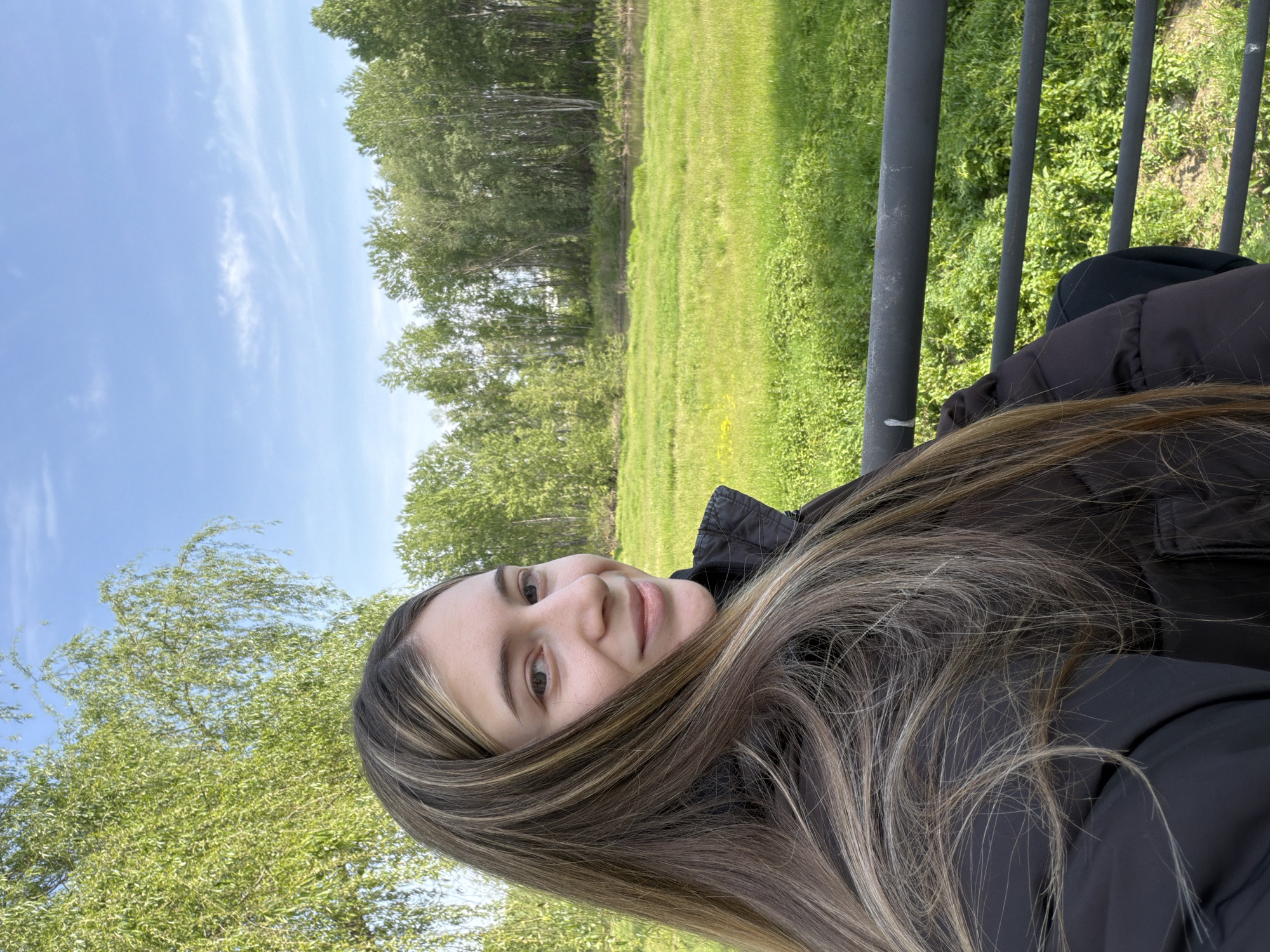 An outdoor portrait features a young woman with long, straight hair, styled with brown and lighter blonde highlights, visible from the chest up. She is positioned towards the left of the frame, looking directly at the camera with a gentle, calm smile. She wears a dark, possibly black or dark brown, jacket.
The background depicts a lush green field or meadow stretching into the distance, bordered by a dense line of deciduous trees. Above the trees, a clear blue sky with a few wispy white clouds is visible. To the right, a dark gray metal fence or railing is partially visible, suggesting she might be leaning against it or standing near a boundary.
The scene is brightly lit by natural sunlight, indicating a clear and sunny day, likely morning or afternoon. The vibrant greenery and blue sky convey pleasant weather. The woman is posing for a photograph. There is no visible text in the image. The setting appears to be a natural, open environment, consistent with a park or countryside area in or around Plovdiv, Bulgaria.