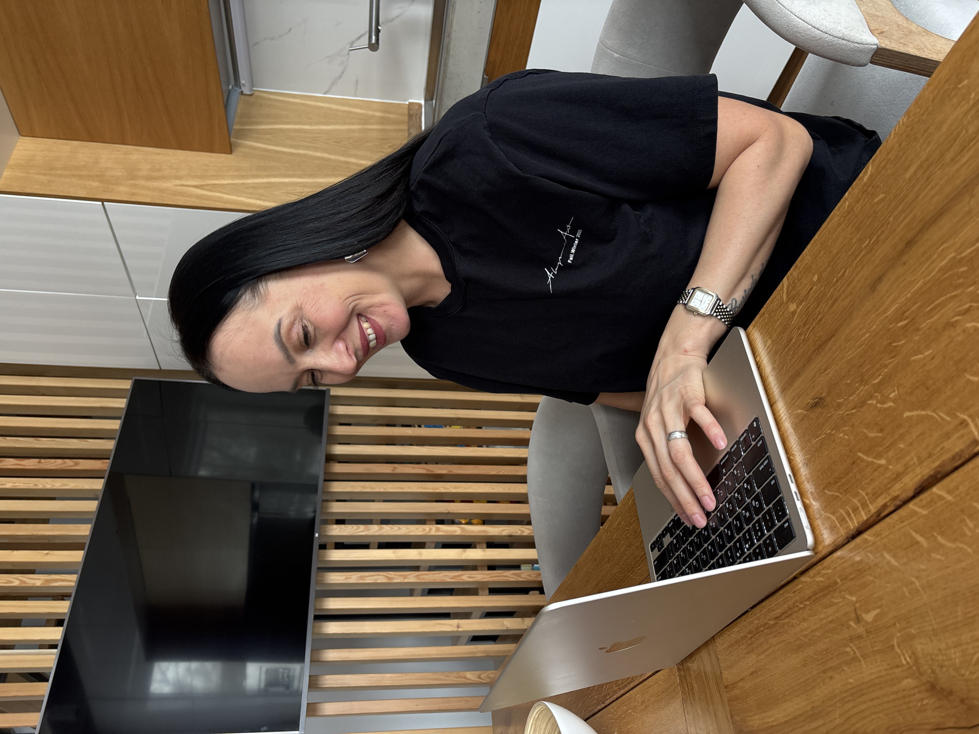 This image depicts an indoor scene in the city of Chaikovsky, Russia, featuring a woman seated at a wooden table, interacting with a laptop. The woman, with long dark hair, is positioned from the chest up, looking downwards and to her left with a smile. She is wearing a black long-sleeved shirt with white text on the chest that reads "Always Aru Fall.Winter 2023." A silver-colored ring is on her left ring finger, and a rectangular silver-colored watch with a mesh band is on her left wrist, where a tattoo is also visible on her forearm. She has geometric silver-colored earrings. Her right hand rests on the keyboard of an open silver-colored Apple MacBook, which is placed on a light brown wooden table with a distinct grain pattern. A light-colored bowl, possibly bamboo, sits on the table to the left of the laptop. In the background, behind the woman, a large, wall-mounted flat-screen television is visible, set against a decorative vertical wooden slat partition. Reflections of a window are present on the television screen. To the right of the woman, a modern kitchen area is visible, featuring white and light brown wooden cabinets, a light-colored countertop or backsplash, and a stainless steel sink faucet. Two light grey upholstered chairs with wooden frames are partially visible around the table.