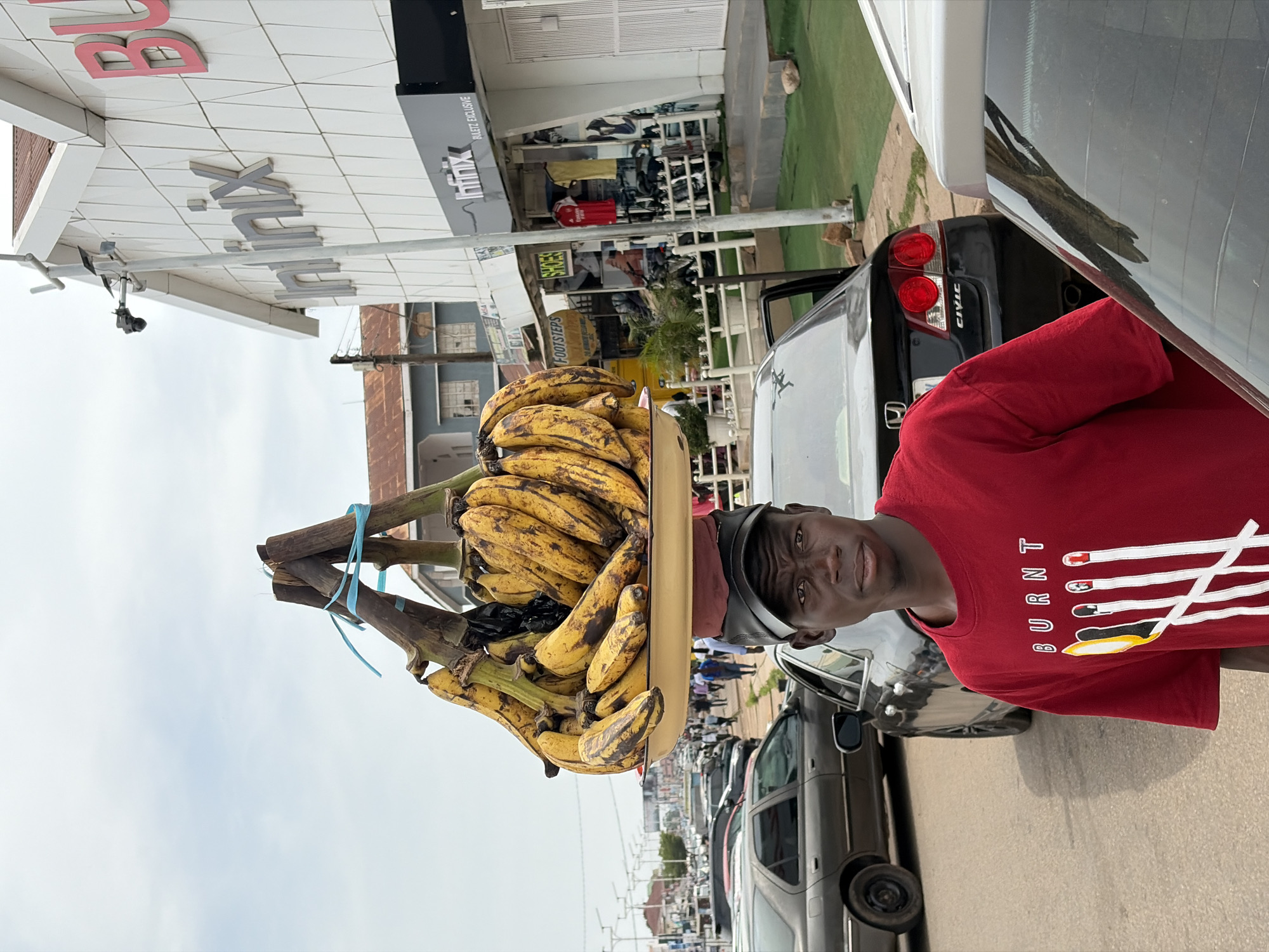 A young man, likely in his late teens or early twenties, stands in the foreground, looking directly at the camera. He is wearing a red t-shirt with white lettering that appears to spell "BURNT" and features a graphic of lit matches. On his head, he balances a large tray filled with bananas. The tray is supported by a wooden structure and secured with blue ties.
Behind the man, a busy street scene unfolds. Several cars are visible, including a dark sedan on the left and a black Honda Civic to his right. The man is positioned near the roadside, which appears to be a paved street with a curb.
In the background, buildings with modern storefronts are present, including one with prominent "infinix" signage. The architecture suggests an urban environment. The sky is overcast. The overall impression is of a market or commercial area in a Nigerian city. The image's metadata indicates the location is Jos, Nigeria.