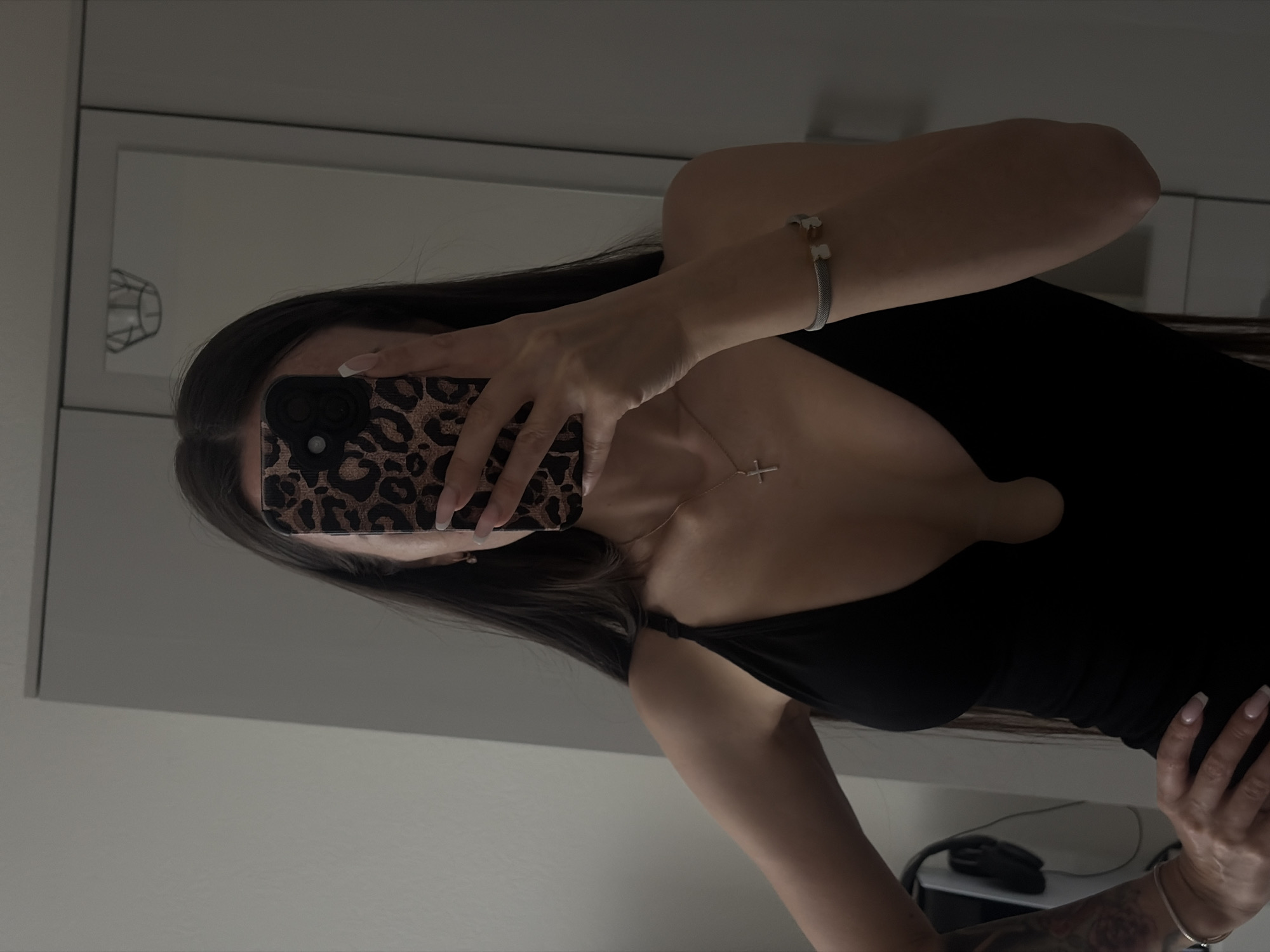 A woman holds a leopard-print phone case in front of her face, obscuring it. She is wearing a black top with a low neckline and a silver necklace with a cross pendant. Her left wrist has a silver bracelet. She appears to be taking a selfie in a mirror, with a blurred image of a light fixture visible in the background. Her long, dark hair flows down her shoulder. Part of a desk and a computer mouse are visible in the lower right corner. The location is Unknown, Unknown.