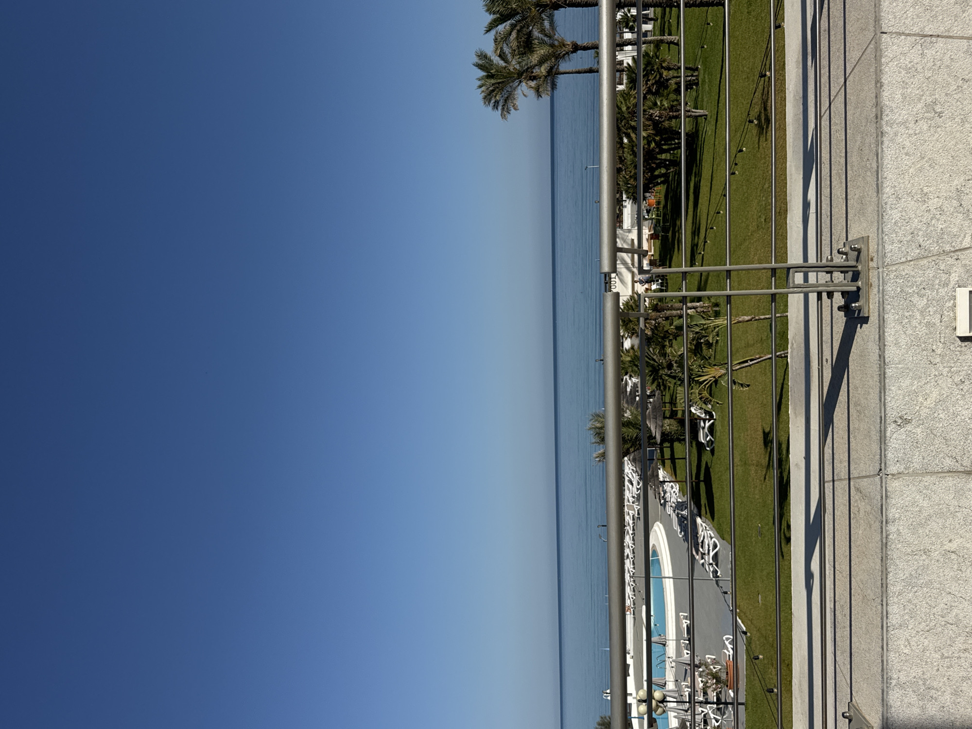 The image shows a view from a balcony overlooking a resort area with a swimming pool and the sea under a clear blue sky. In the foreground, a metal railing with multiple horizontal bars runs across the frame, attached to a concrete structure. Beyond the railing, a green lawn is visible, dotted with rows of empty lounge chairs and umbrellas. A swimming pool with blue water is situated to the left, also surrounded by lounge chairs. Palm trees are scattered throughout the resort area, with a cluster of taller palm trees visible on the right side of the frame, near the sea. The sea itself is calm and stretches to the horizon, meeting the sky in a distinct line. The scene appears to be a sunny day at a coastal resort, likely in Isola delle Femmine, Italy, given the Mediterranean vegetation and the style of the resort. No people are actively engaged in any activity, though a few figures are visible in the distance near the pool area. The overall atmosphere is tranquil and depicts a vacation setting.