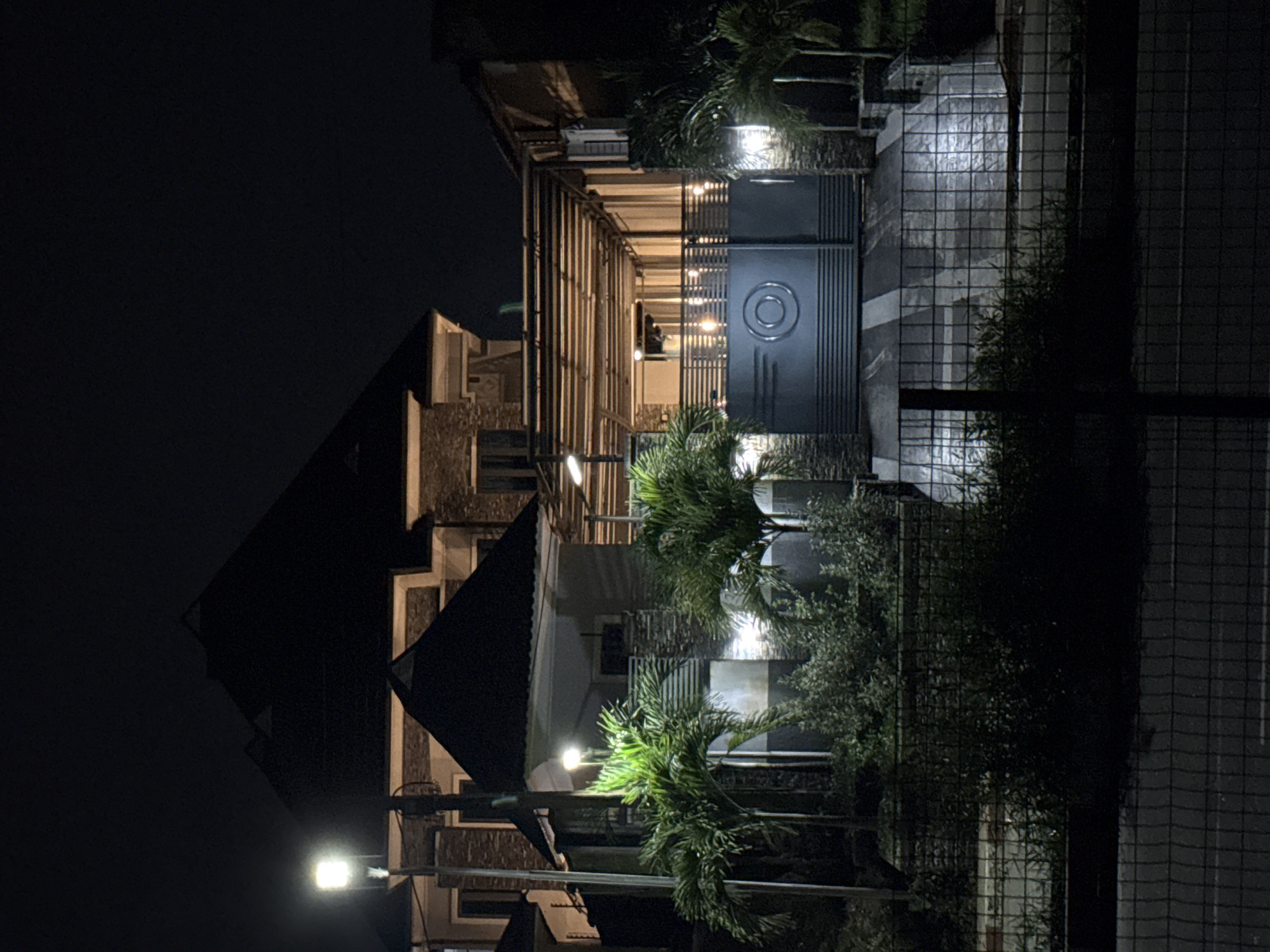 The image depicts a nighttime scene in Uyo, Nigeria, featuring a modern house with distinctive architectural elements. The house has a multi-tiered, dark-colored roof and stone-clad exterior walls. A prominent feature is a large, dark-colored metal gate with a circular emblem, suggesting a sense of security or exclusivity. In front of the gate, a driveway is visible, with striped patterns creating visual interest. Palm trees and lush green foliage surround the property, adding a tropical feel to the environment. The house is illuminated by artificial lighting, including streetlights and lights integrated into the building's design, casting a warm glow on the surroundings. The overall impression is one of a well-maintained and illuminated residence in a suburban or residential area of Uyo.