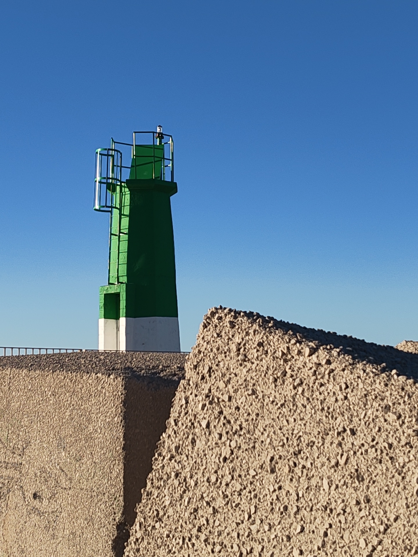 The image depicts a green and white lighthouse in Dénia, Spain, standing atop a concrete structure alongside a rough, pebbled breakwater. The sky is clear and blue. The lighthouse is a simple, cylindrical structure with a small, enclosed top and metal railings. The lower portion of the lighthouse is painted white, while the upper portion is green. The concrete structure it sits on is textured, and a railing is visible. The breakwater in the foreground is composed of irregularly shaped, light-colored stones. There are no people visible in the frame.