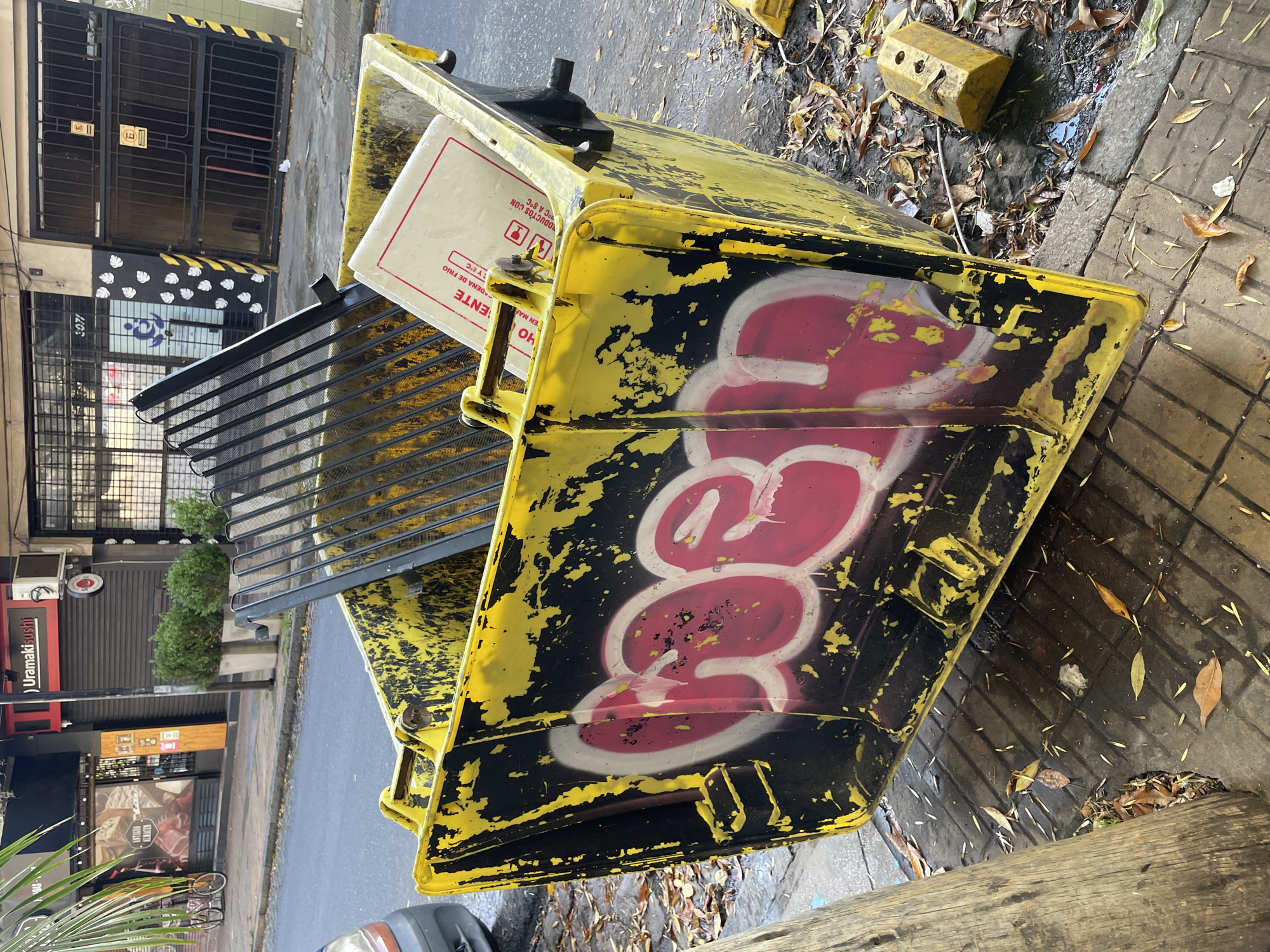 The image shows a yellow and black, heavily graffitied dumpster sitting on a street in Olivos, Argentina. The dumpster is open and contains a white box labeled "ALMACEN ANTIGUO". The box also has instructions in Spanish. Behind the dumpster is a wrought-iron gate in front of a building with the number "3071" visible. Across the street is a restaurant named "Uramaki sushi". The street is wet and littered with leaves.