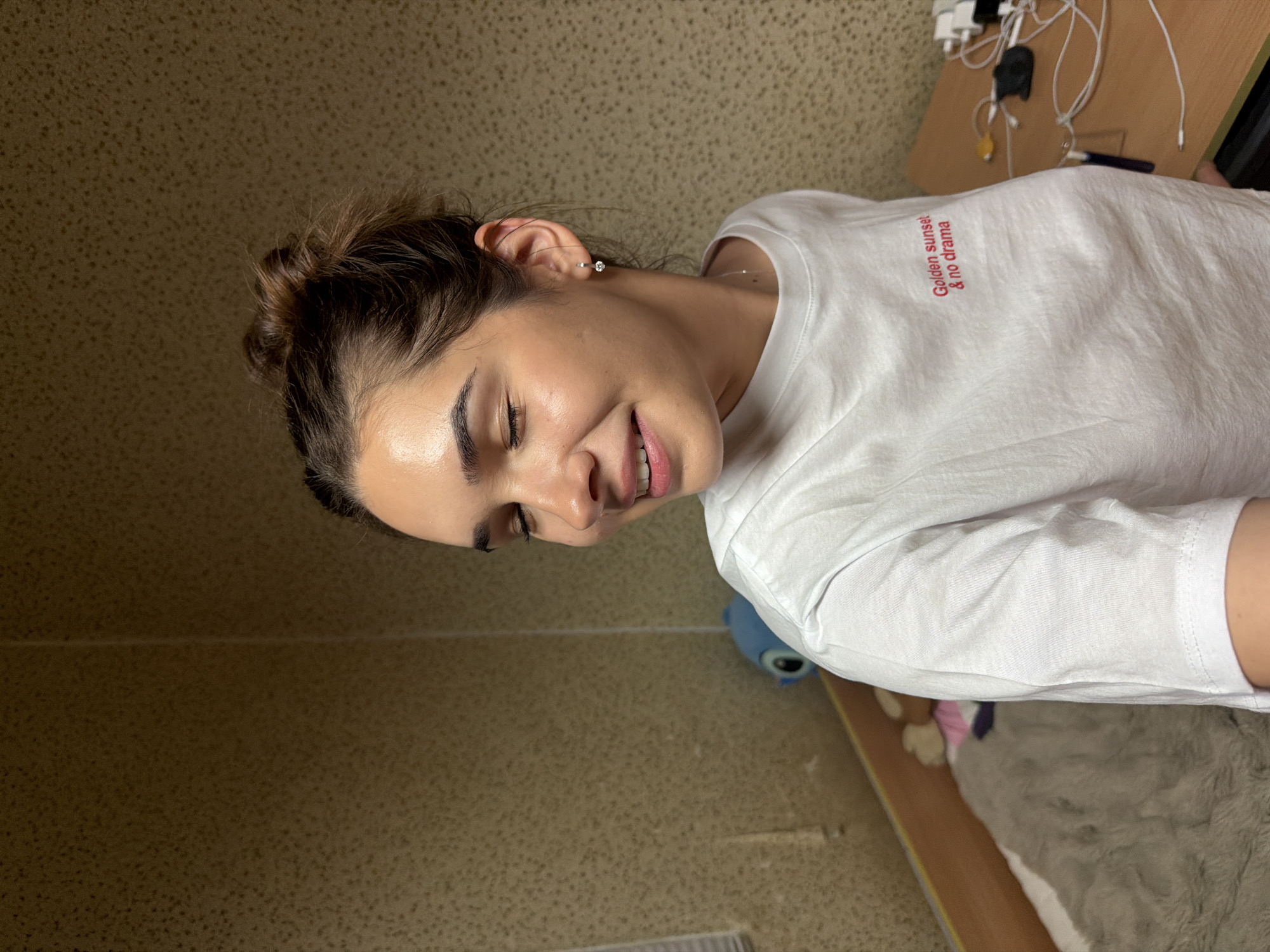A woman with dark hair pulled back in a bun is wearing a white t-shirt with red text that reads "Golden Sunset & no drama". She is smiling with her eyes closed slightly. The background shows a patterned beige wall. A desk with wires and devices is visible on the right, along with a stuffed animal and a blue object. The scene is indoors and appears to be in a personal residence. There is no information available to conclude this image was taken in Sofia, Bulgaria.