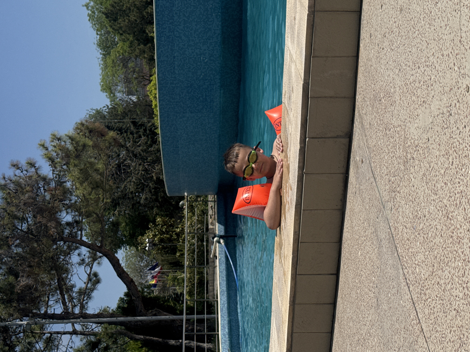 A young boy with goggles and orange arm floaties is at the edge of a blue-tiled swimming pool. The pool is bordered by a tile wall and a concrete patio. A tree and some fencing are visible beyond the pool. This photo was likely taken in Yevpatoriya, Ukraine.