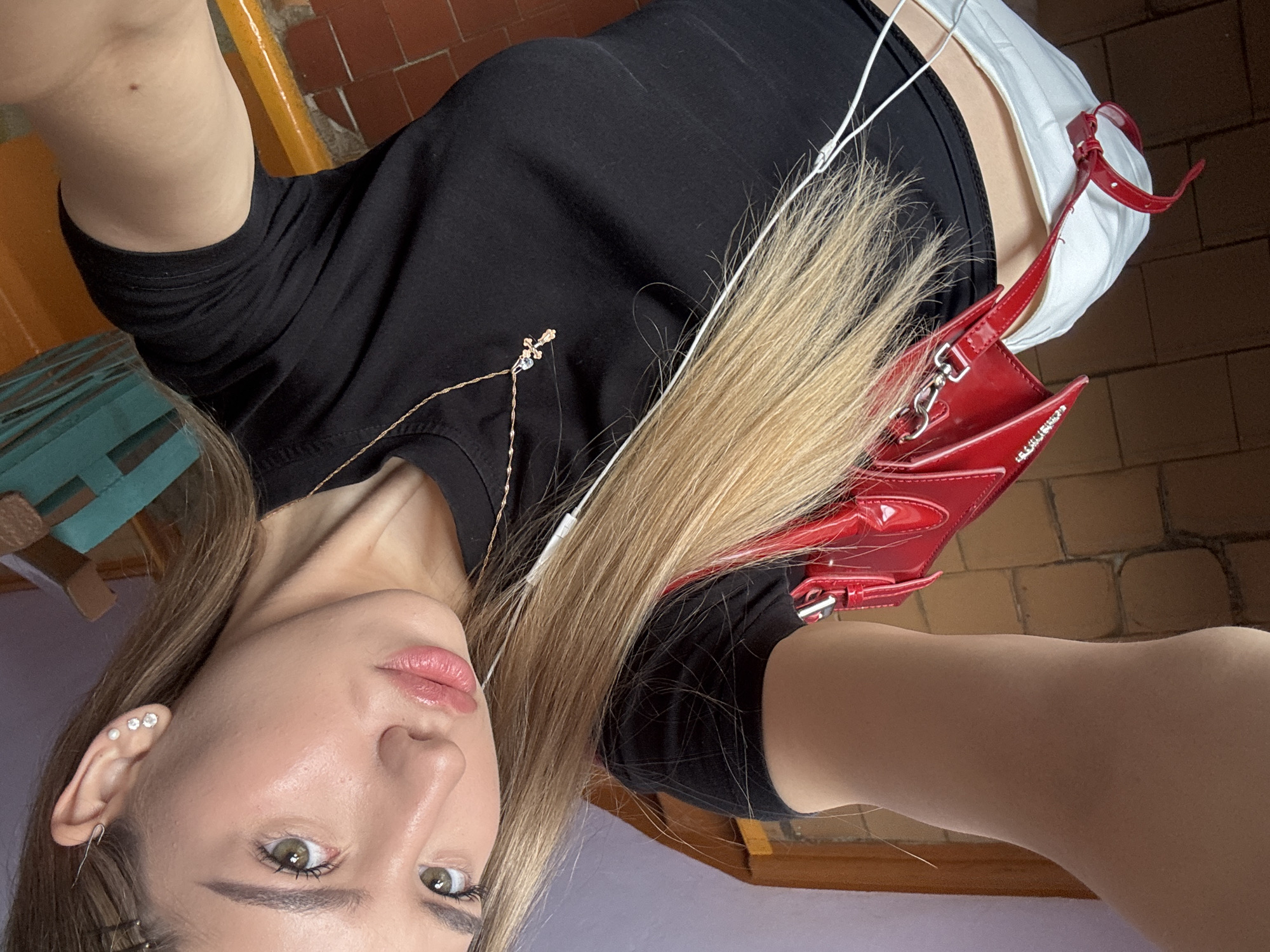 A woman takes a selfie in an indoor location with tiled floors and brick walls. She is wearing a black top, white pants, and a red handbag. She has blonde hair and is wearing earrings and a necklace. The location is assumed to be in the city of Noviy, Russia.