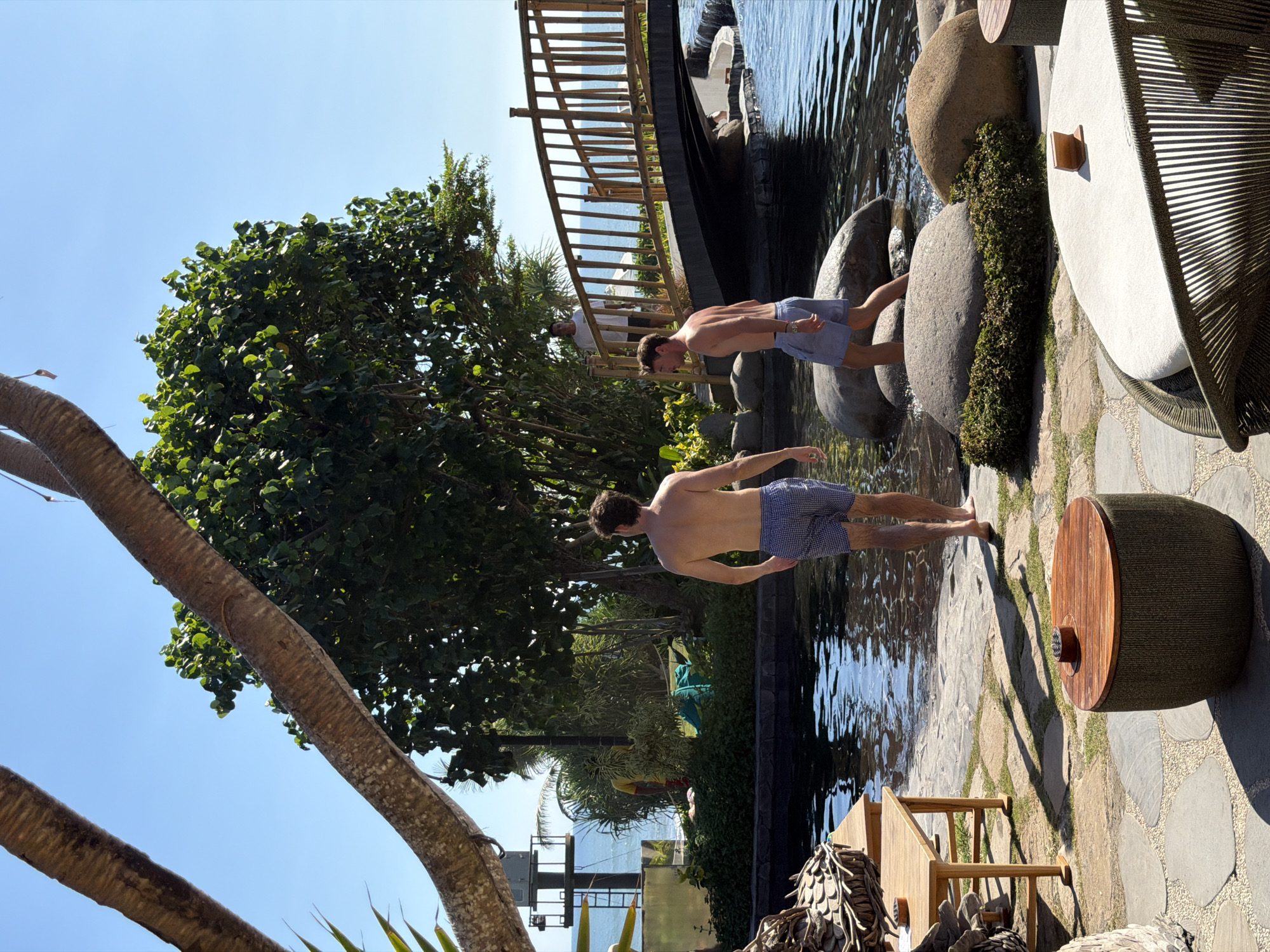 Two shirtless men, wearing swim trunks, are walking by a body of water. The man on the left, with dark hair, is facing away from the camera and towards the water. He is wearing blue and white checkered swim trunks. The man on the right, also with dark hair, is facing towards the water with his back partially to the camera. He is wearing light blue swim trunks. They are in an outdoor environment with lush green trees, large rocks, and a wooden bridge in the background. The water appears to be a pond or a shallow pool, with reflections of the sky and trees on its surface. There are also pieces of outdoor furniture visible, including a wooden table and chairs. The scene suggests a resort or a recreational area, possibly in Kediri, Indonesia, given the tropical vegetation and the warm, sunny weather.