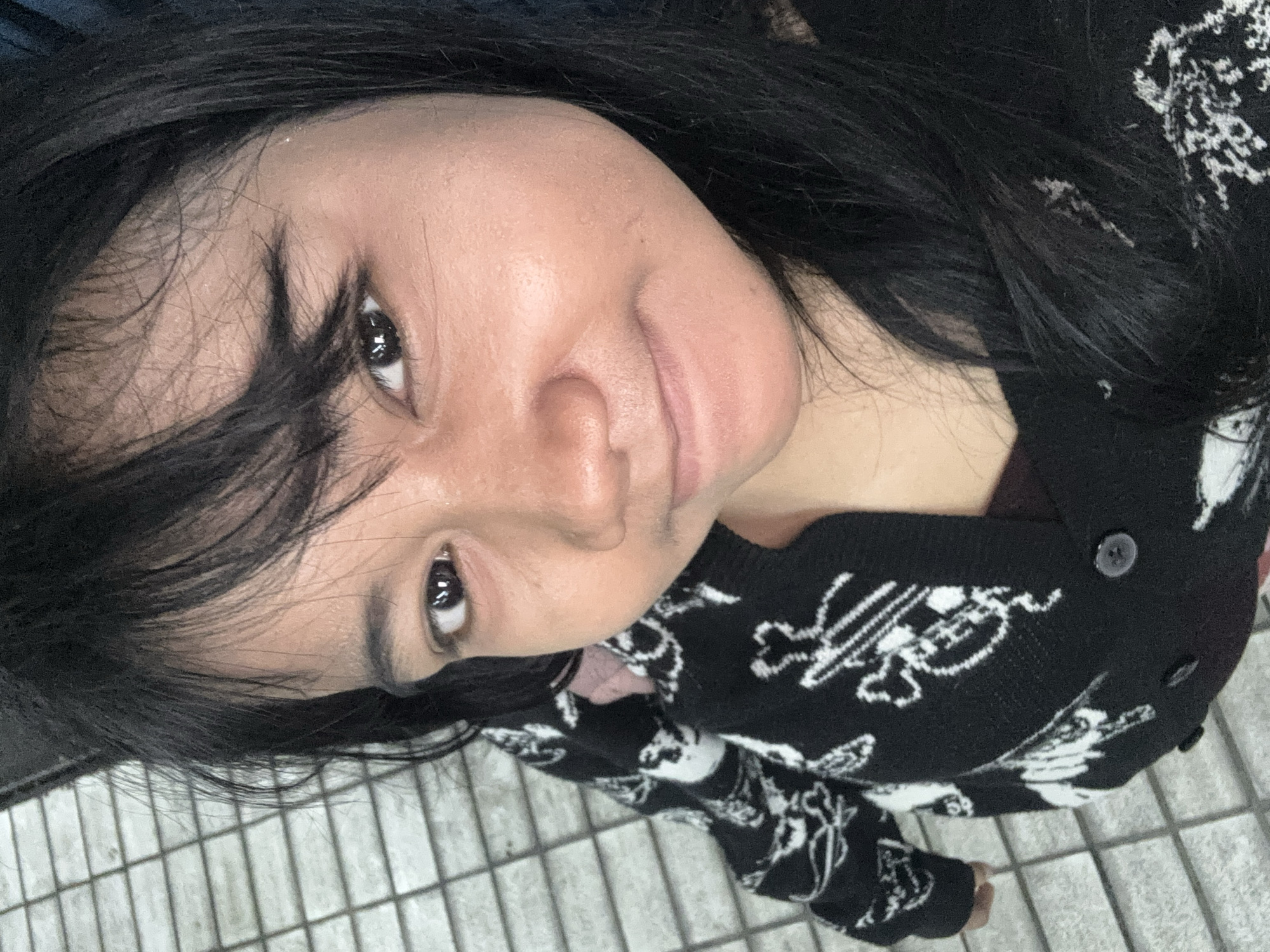 A selfie of a young East Asian woman with dark hair and brown eyes is taken from a high angle, looking down at the camera. She is wearing a black cardigan with a white skull and crossbones pattern over a maroon shirt. The background shows a gray tiled floor, possibly a sidewalk or walkway. The lighting suggests it is daytime, possibly outdoors. Her facial expression is a slight smirk.