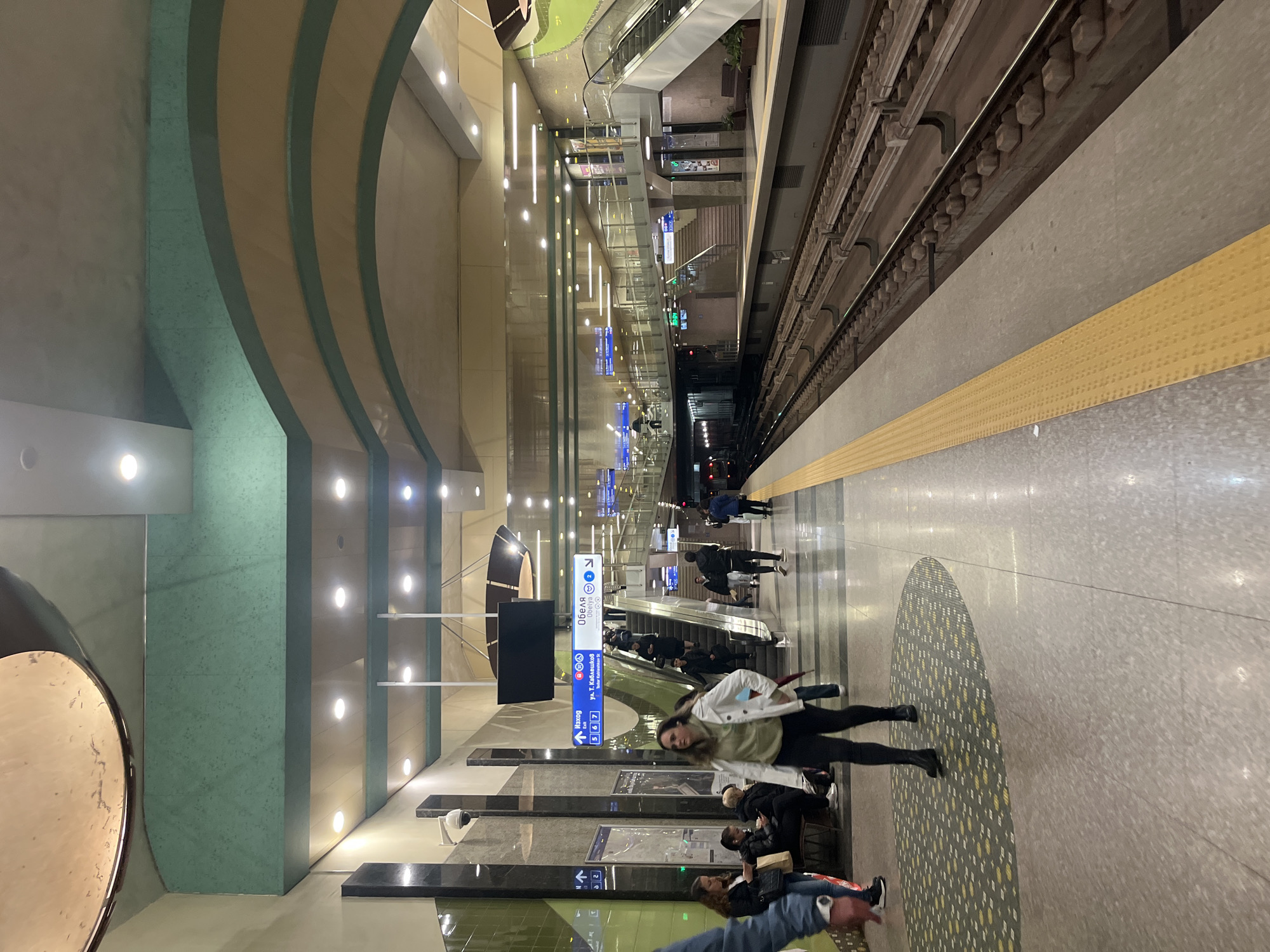 This media file depicts a modern and brightly lit underground metro station platform, identified as being in Sofia, Bulgaria. The station features contemporary architectural design with a distinctive ceiling and wall treatment comprising curved, alternating panels of light beige and a muted teal-green. Artificial lighting, including recessed spotlights and vertical strip lights, illuminates the entire space, suggesting it could be any time of day or night.
On the right side of the image, subway tracks are visible, running alongside the platform, which is marked by a yellow tactile strip for safety. In the background, escalators and stairs lead to different levels of the station. Several people are present on the platform; in the foreground, a woman in a white jacket and dark pants walks towards the right. Further back, other individuals are seen sitting on benches or standing, appearing to wait for a train or navigate the station. The floor is a light-colored terrazzo with a large, circular patterned inlay featuring grey, white, and yellow elements.
A prominent blue and white directional sign is visible, displaying "ОБЕЛЯ" (OBELYA) with metro line symbols and the Olympic rings. Below this, "Sofia" with an arrow, "Ул. Т. Каблешков" (Tsar Kableshkov St), and metro lines 5, 6, and 7 are listed. The word "Изход" (Exit) is also clearly visible on the sign. A circular, metallic decorative fixture is mounted on the left wall. The atmosphere is functional and orderly, typical of a public transportation hub.