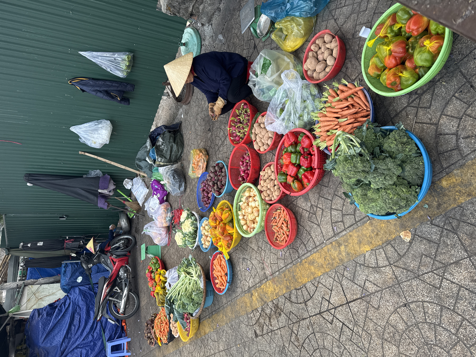 A street vendor sits crouched next to a collection of produce baskets in a marketplace in Da Lat, Vietnam. The vendor, wearing a traditional conical hat and a dark blue jacket, sits on the concrete sidewalk next to a green metal wall. Baskets filled with a variety of vegetables – including potatoes, carrots, peppers, onions, broccoli, cauliflower, and radishes – are arranged in a line along the sidewalk. The ground is marked with yellow lines on a patterned, concrete sidewalk. A motorcycle is parked behind the vendor, partially covered by a blue tarp. Several bags and other miscellaneous items are scattered around the area. A weighing scale sits nearby. The image suggests a typical local market scene in the city.