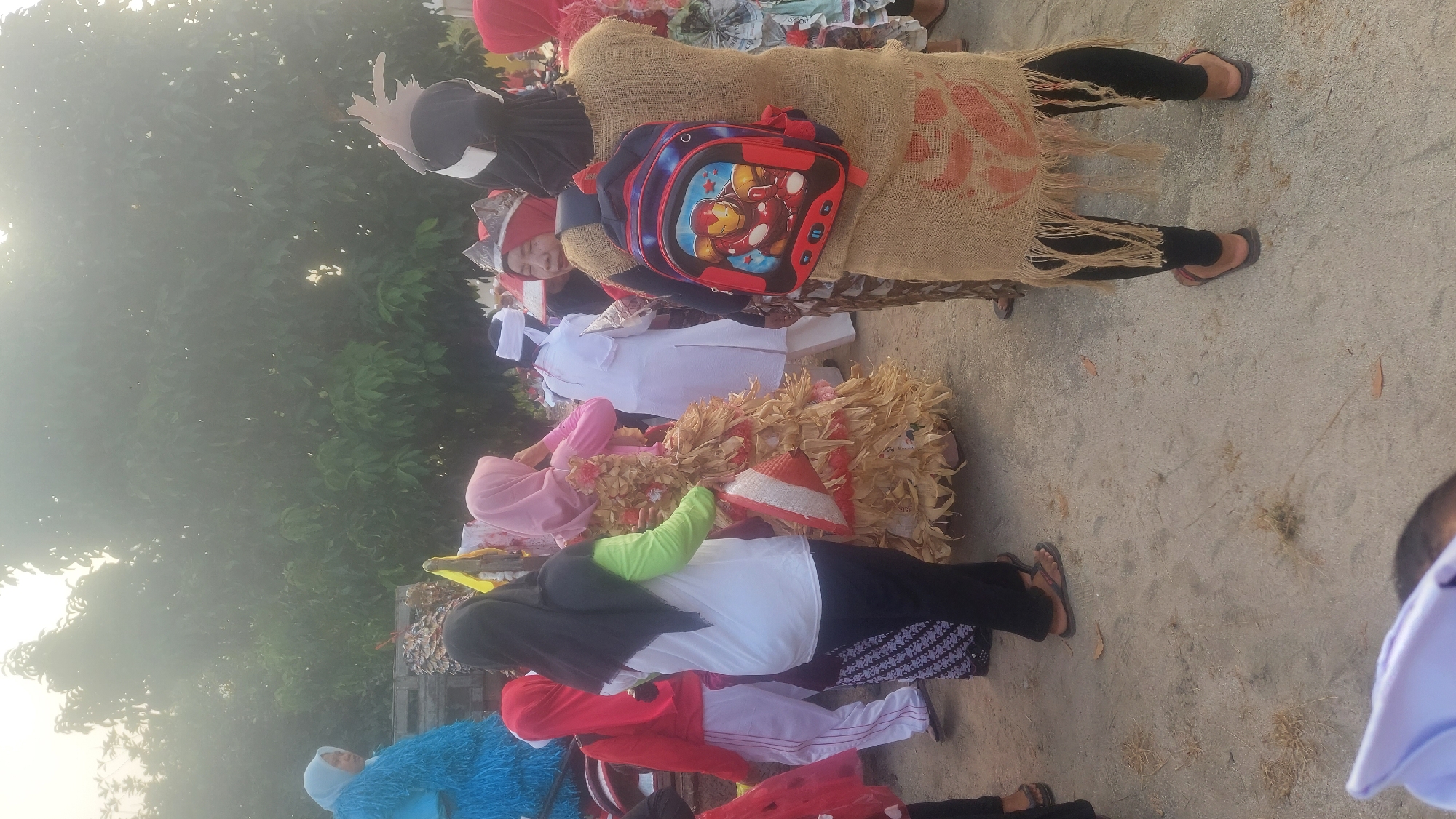 In a dusty outdoor area, possibly a street or field, a group of people are gathered, many in costumes. The ground is a mix of dirt and sand, with some dry vegetation scattered about. In the background, a dense canopy of green trees creates a natural backdrop, with the sky above appearing hazy, possibly due to fog or sunlight.
Several individuals are dressed in colorful attire. On the far left, a person wears a large, fuzzy blue costume. Next to them, a person in a red jacket and white pants with red stripes stands. A woman in a black hijab and green shirt, black pants, and sandals is in the center, holding a conical object with red and white stripes. To her right, a person in a pink hijab wears a voluminous dress made of dried, straw-like material. Further right, someone in a white outfit and white head covering is visible.
A prominent figure in the foreground on the right is wearing a burlap-like sack costume draped over black leggings and red-brown sandals. They are also carrying a red backpack adorned with an image of Iron Man. The overall scene suggests an outdoor event or parade, possibly a cultural celebration or festival, taking place in Reno Basuki, Indonesia.