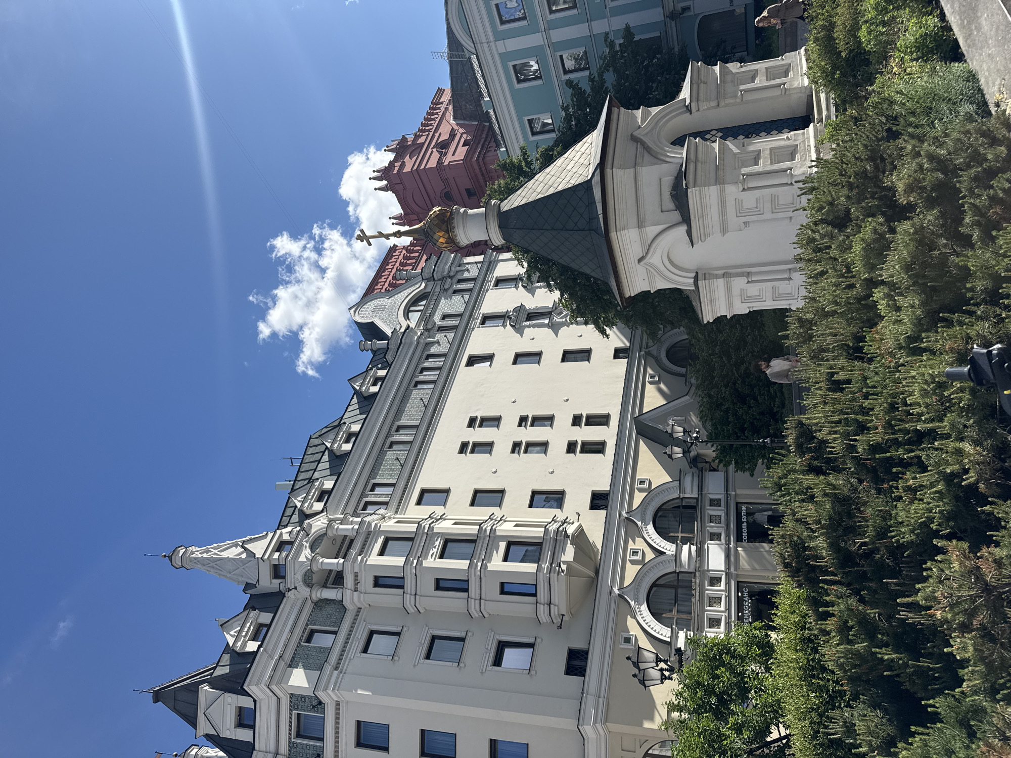 The image shows a street view in Moscow, Russia, featuring the Metropol Hotel. The hotel, predominantly white with architectural details like ornate windows, turrets, and a chapel with a golden dome, dominates the scene. Other buildings with distinctive architectural styles are visible in the background, including a red structure and a light blue building. The sky is clear blue with some clouds, and sunlight illuminates the scene. Trees and bushes are present in the foreground, partially obscuring the lower part of the buildings. Two people are visible in the distance. Street lamps line the building's facade.
