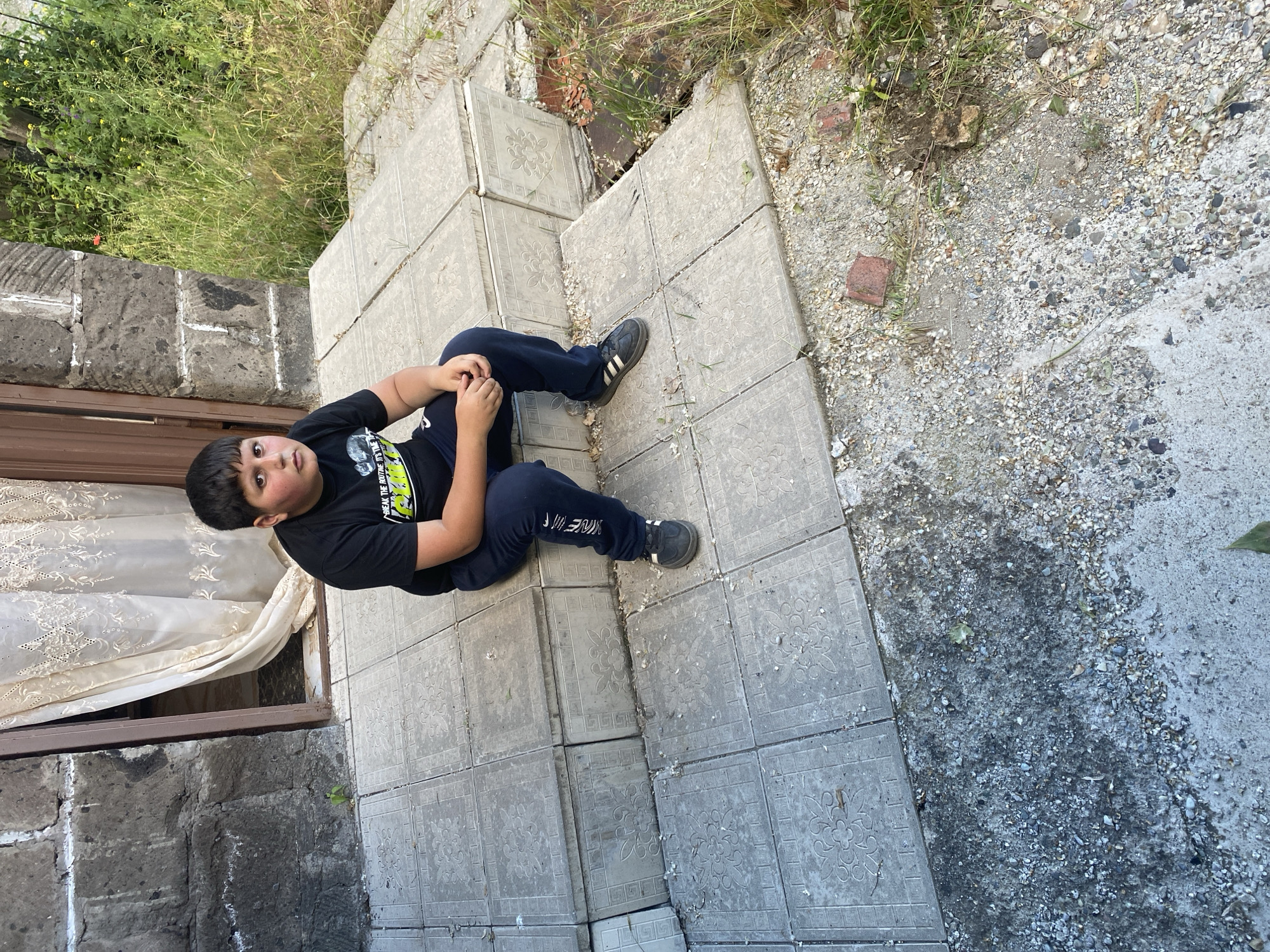 A young boy with dark hair and brown eyes sits on a tiled outdoor staircase. He is wearing a black t-shirt with a graphic design and navy blue track pants. The boy is looking directly at the camera with a neutral expression. He is sitting with his legs crossed, and his hands are resting in his lap. The staircase is made of grey tiles with a floral pattern. To the left of the staircase is a stone wall. To the right of the staircase is overgrown green grass and vegetation. The scene appears to be in the city of Akhurik, Armenia.