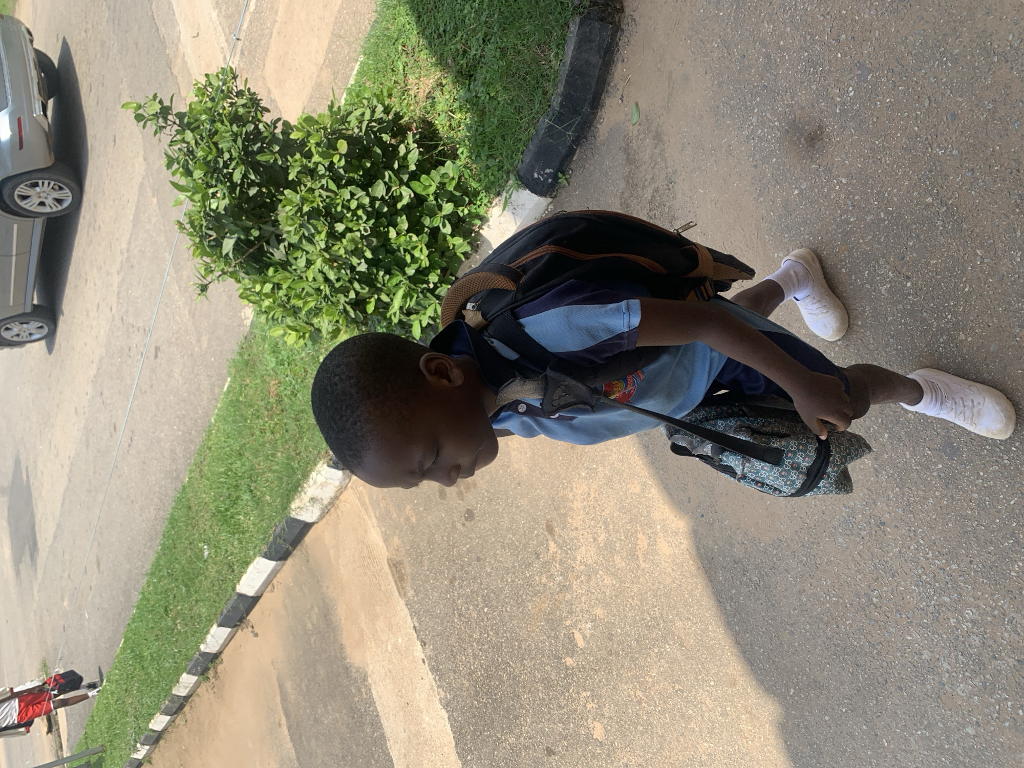 The image depicts a young Black male child walking on an asphalt path. He is wearing a light blue polo shirt with a crest on the left side, navy blue shorts, white socks, and white athletic shoes. The child is carrying a black backpack with brown straps and a smaller, patterned bag in his right hand. The environment appears to be outdoors, with a patch of green grass and a low, black and white curb to the left of the path. In the background, a light-colored SUV is visible on a paved surface, and a dense green shrub is situated between the path and the road. The lighting suggests it is daytime, with shadows cast on the asphalt. The scene is likely a public space within Abuja, Nigeria, possibly near a school or residential area.