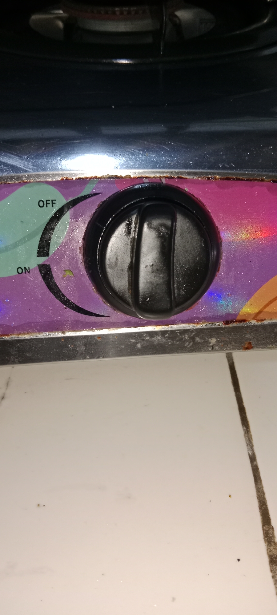 The image shows a close-up of a gas stove knob, indicating the "ON" and "OFF" settings. The knob is black and appears dirty. The stove has a metallic surface with a colorful, possibly holographic, pattern. The background consists of light-colored tiles. The scene is likely an indoor domestic setting, common in areas such as Subang, Indonesia, where gas stoves are a primary cooking appliance. There are no people or actions visible in the image. The lighting suggests an artificial light source, possibly from above. The overall condition of the knob and surrounding surface indicates regular use and potential lack of recent cleaning.