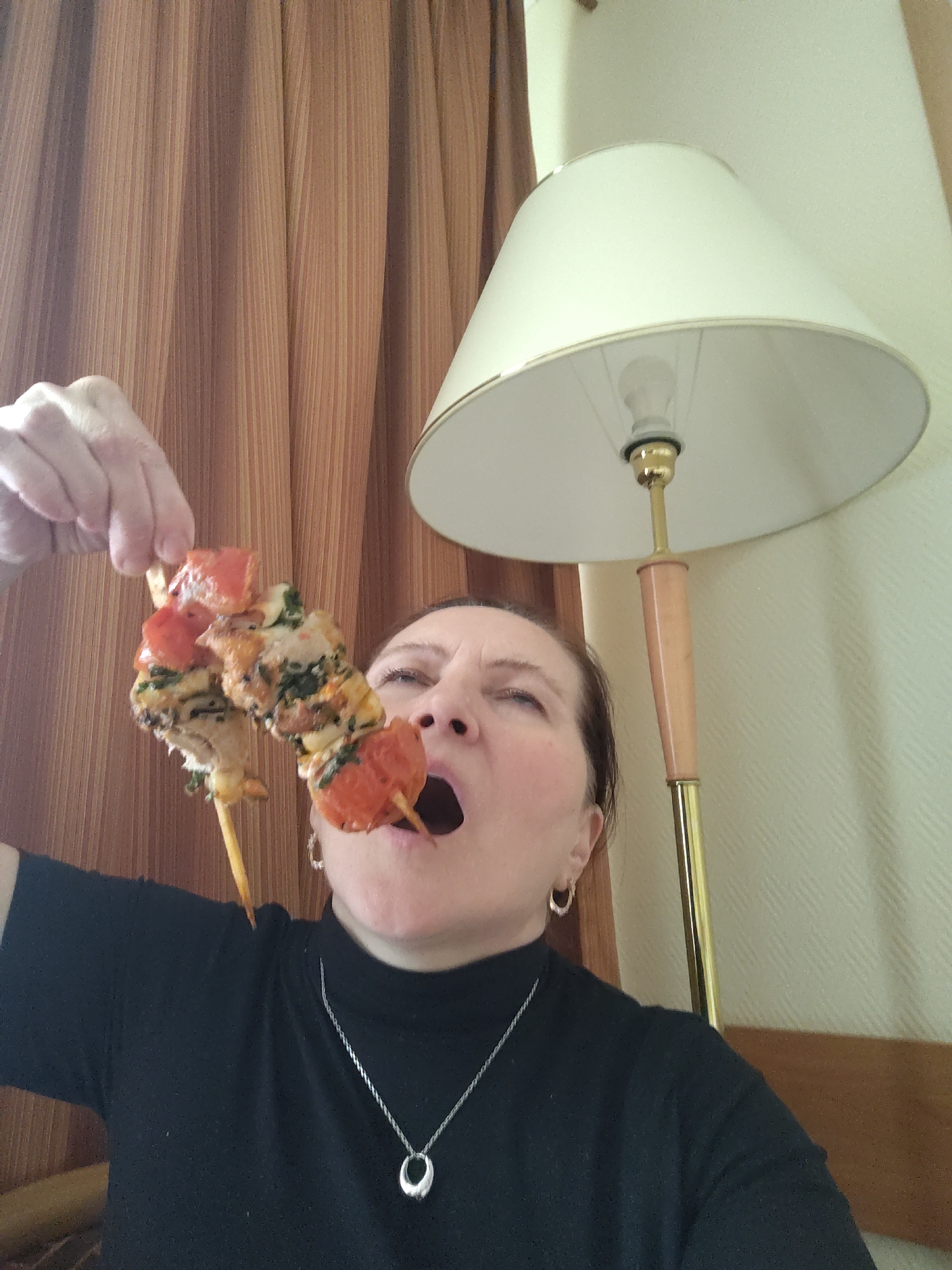 This indoor photograph, taken in Moscow, Russia, captures a woman attempting to eat two skewers of kebabs. The woman, appearing middle-aged with dark hair, is looking upwards with her mouth wide open and eyes partially closed, conveying a sense of humor or exaggerated effort as she prepares to bite. She wears a black high-neck top, a silver pendant necklace with an oval-shaped charm, and small hoop earrings. Her left hand, positioned overhead, holds the two wooden skewers which are laden with cubed chicken or meat, bright red tomato pieces, and green herbs.
The background features brown, vertically-striped curtains on the left side of the frame. On the right, a cream-colored wall displays a subtle, repeating diagonal or diamond texture. A prominent floor lamp stands on the right, equipped with a large, cream-colored conical shade. The lamp's bulb is visible inside, attached to a golden fixture and a light wooden stem that extends down to a golden base. The lighting suggests an indoor setting, possibly a room in an apartment or hotel. No text is visible in the image.