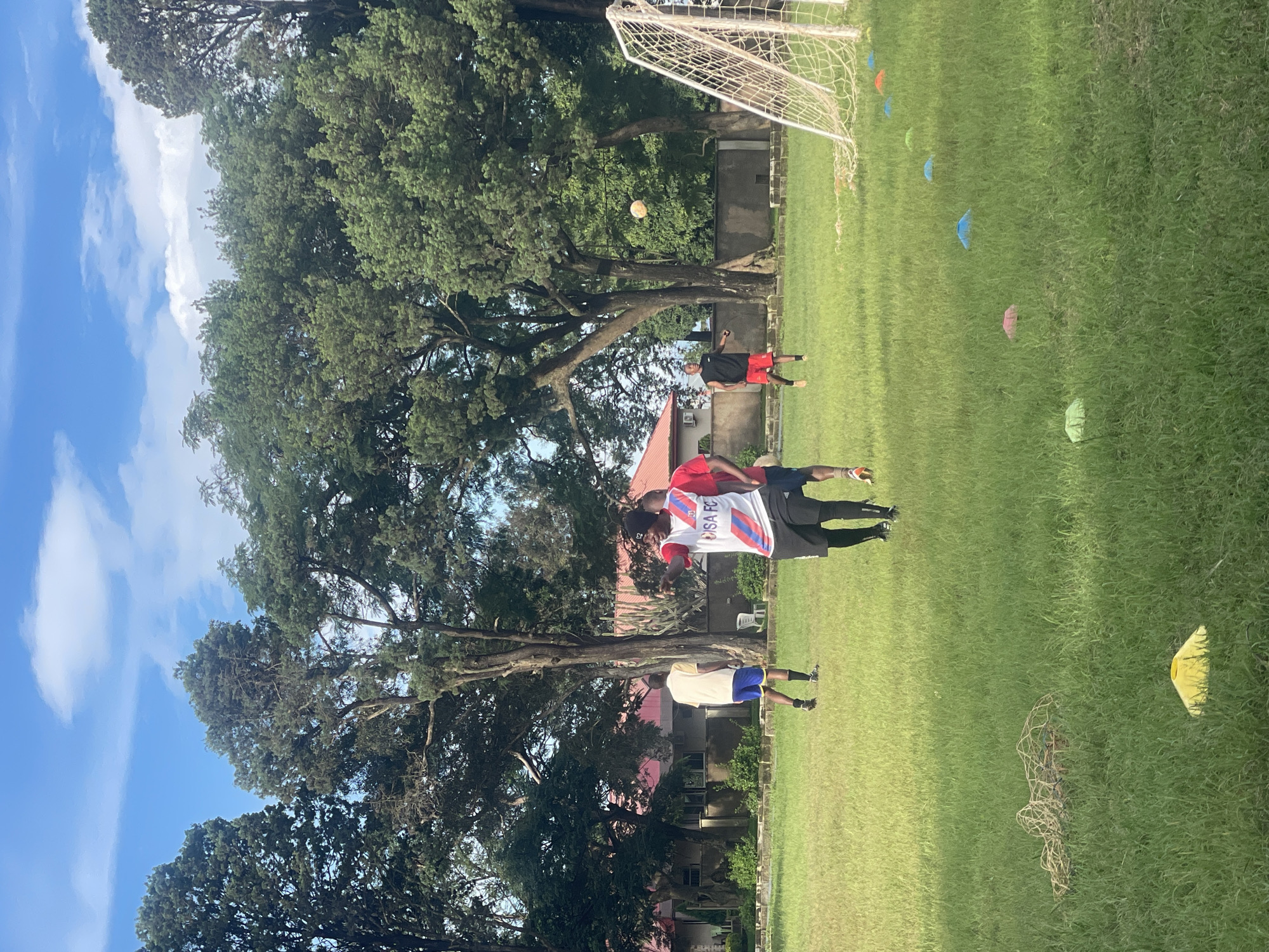 The image depicts a soccer field on a bright, partly cloudy day, likely within the city of Jos, Nigeria. Several individuals are engaged in a soccer practice. In the foreground, a person wearing a white, red, and blue jersey with "DISA FC" printed on it and black shorts, is facing left, pointing with their right hand and appearing to give instructions or directions. To their right, another individual in a red shirt and black shorts is visible from the chest down, also facing left.
Further in the background, on the left side of the frame, a person in a yellow shirt and blue shorts is walking away from the camera. In the center, a person in a black sleeveless shirt and red shorts is kicking a soccer ball which is in mid-air. Another person is partially visible on the far right, wearing a black shirt and black shorts.
The environment consists of a well-maintained green grass field. Several colorful training cones are scattered across the field, with a yellow cone prominent in the lower left, and rows of blue, green, and orange cones leading towards a soccer goal on the right. A deflated soccer net lies on the grass in the middle ground to the left. Beyond the field, there are large, mature trees with dense foliage, and in the distance, a building with a red roof and light-colored walls. The sky is blue with scattered white clouds.