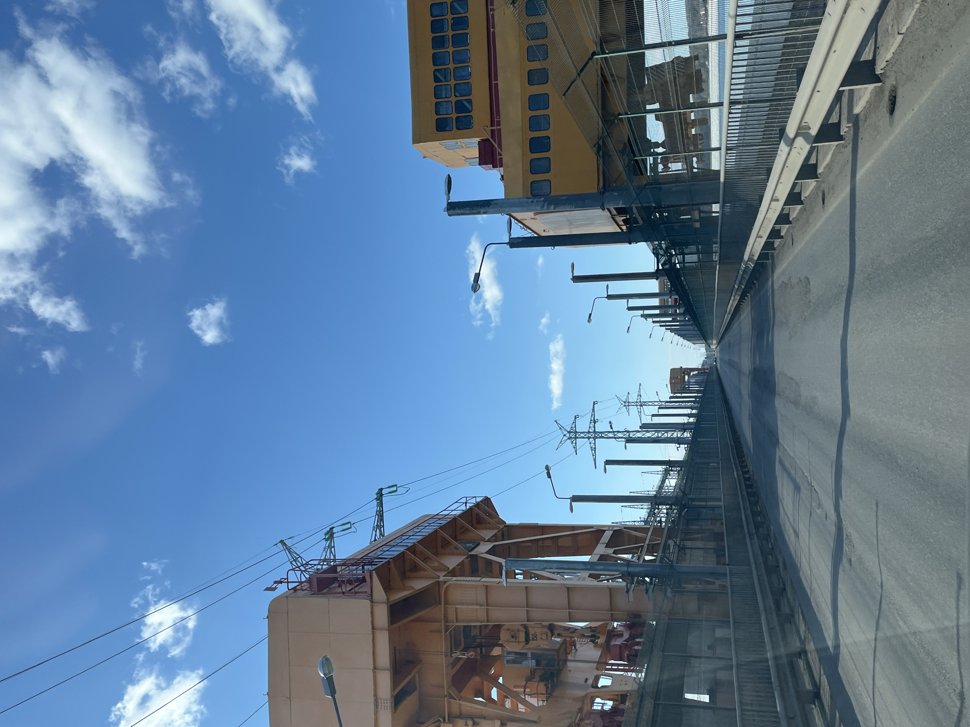 The image shows a view of the Kama Bridge in Chaikovsky, Russia, from a vehicle traveling along the bridge. The bridge is lined with a fence and utility poles. Large, industrial structures, possibly related to power distribution, are situated on either side of the bridge. The sky is blue with scattered white clouds. The road surface is asphalt. The camera's point of view is from inside a vehicle, as indicated by the windshield reflection.