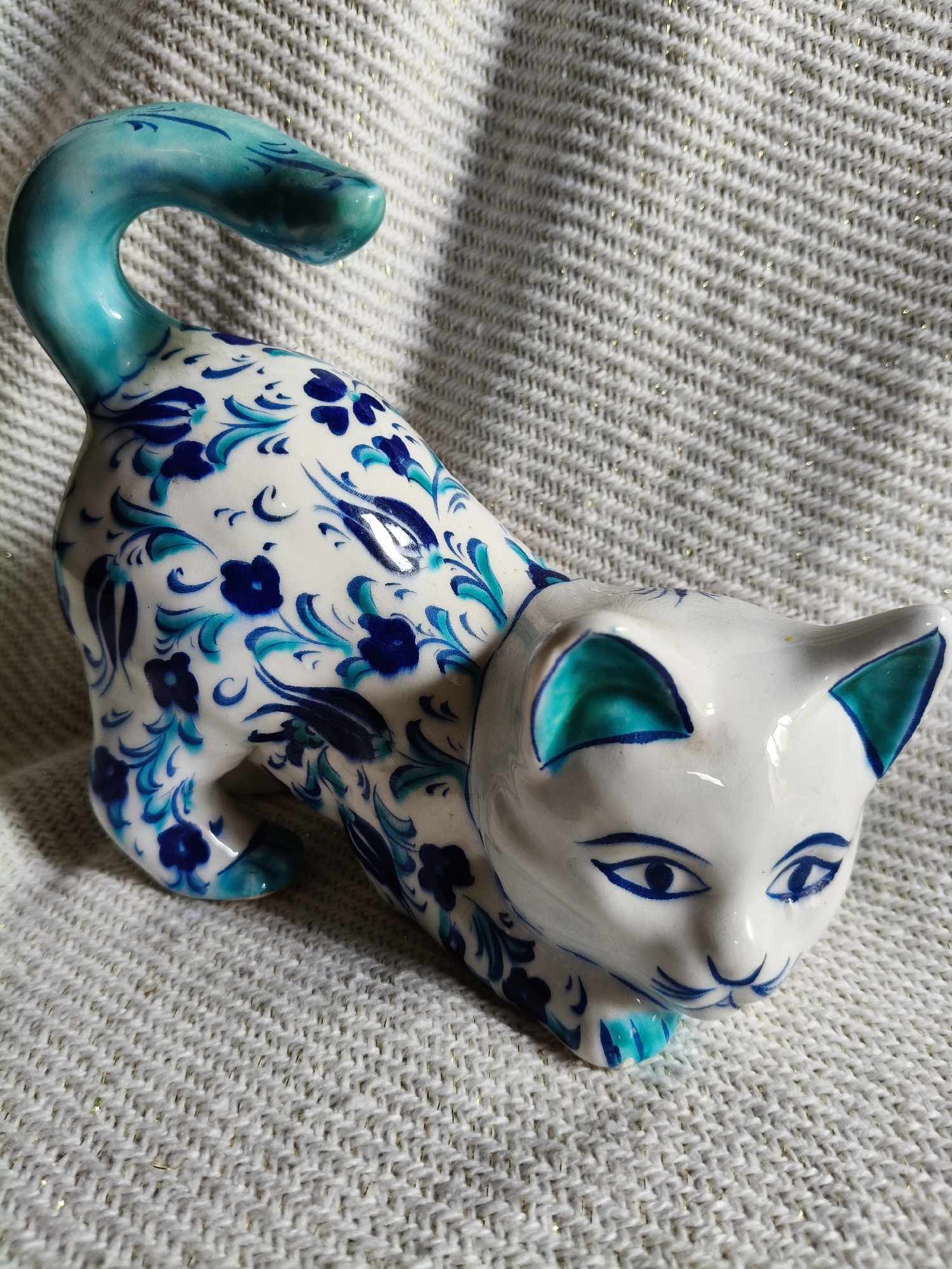A white ceramic cat figurine with blue and teal floral patterns and accents is displayed on a textured white fabric with gold threads. The cat is in a crouched position, facing right, with its tail curled upwards. The figurine has detailed blue eyes, a small nose, and whiskers. The fabric appears to be a blanket or throw. The image does not provide any information about its location in Bigauņciems, Latvia.