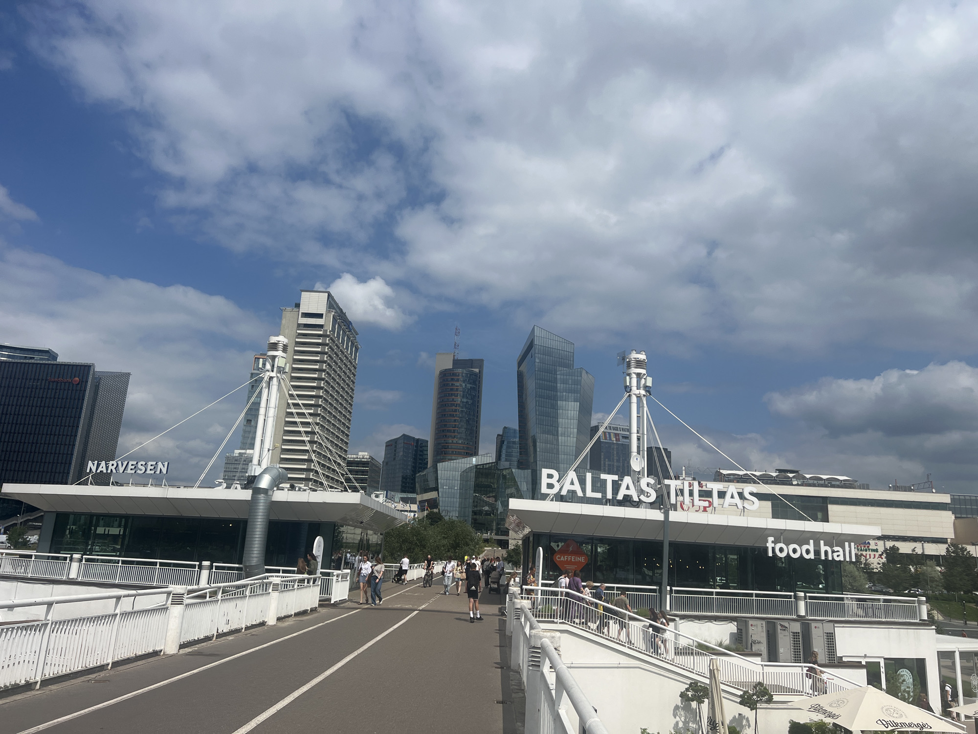 The image depicts a sunny day scene on a pedestrian bridge in Vilnius, Lithuania, with a modern cityscape in the background. The bridge is lined with white railings and features a pathway with markings for walking and cycling. Several people are walking and cycling on the bridge, engaged in various activities.
On the left side of the bridge, a building with the signage "NARVESEN" is visible. To the right, another building prominently displays the signage "BALTAS TILTAS" and "food hall". Several modern skyscrapers and office buildings form the urban skyline in the distance. The sky is partially cloudy, with patches of blue. The overall atmosphere is lively and urban, indicative of a public space in a city.