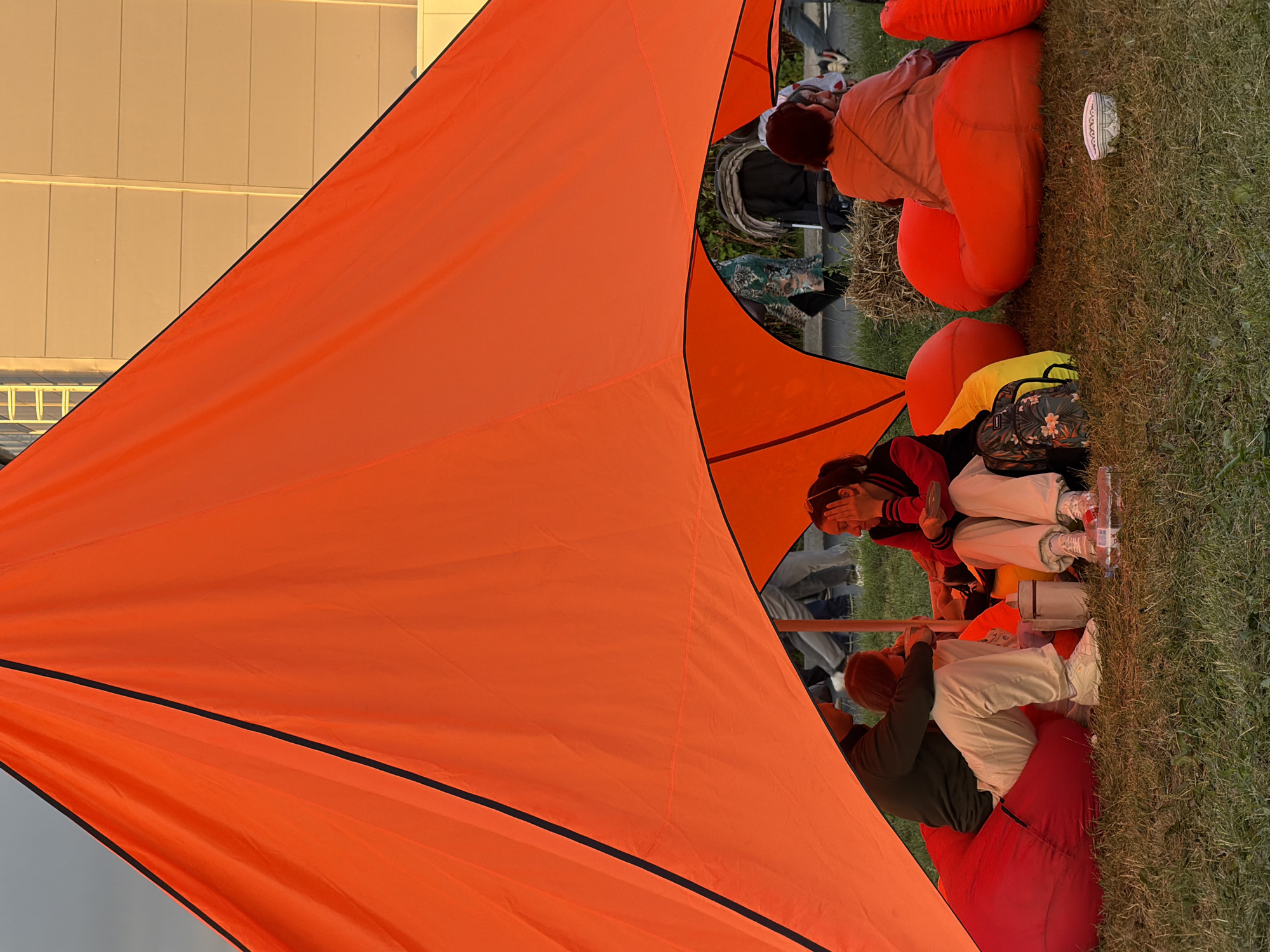 The image depicts an outdoor scene under a large, bright orange canopy. Several people are seated on large, red beanbag chairs on a grassy area. On the left, a person wearing a dark green long-sleeved shirt and white pants sits on a beanbag, holding a white cup. Next to them, another person with blonde hair is also seated. To the right, a person in a red and black jacket and light-colored pants is touching their face. Further to the right, another individual in an orange top is seated, facing away from the camera. In the background, more people are visible, along with a stroller and some hay bales. The environment appears to be a park or an outdoor gathering space, with a building visible in the upper right corner. The lighting suggests it might be late afternoon or early evening.