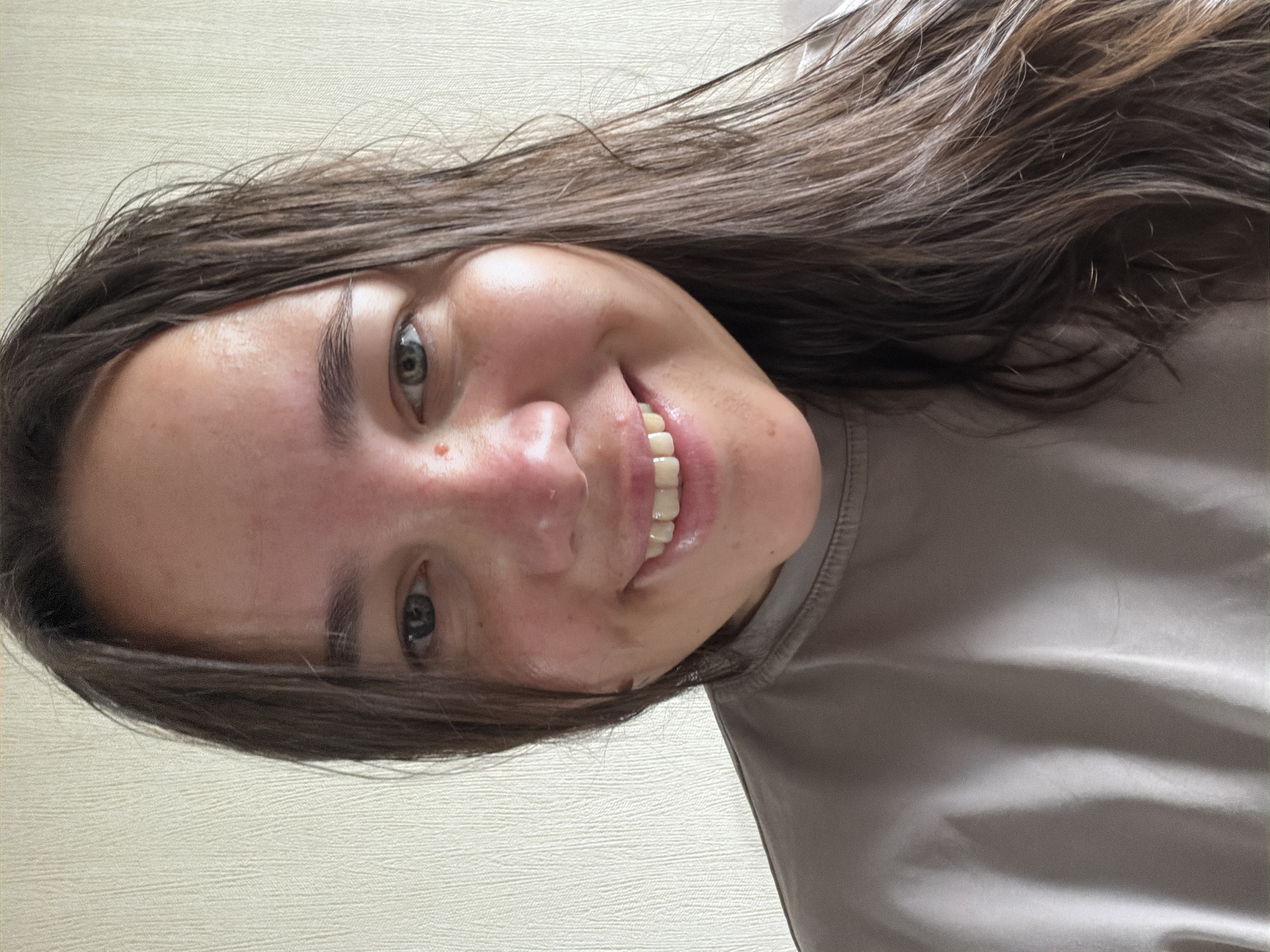 A close-up selfie of a young woman with long, dark brown hair is shown. She has fair skin with some redness on her cheeks and nose, a small mole on her cheek, and bright blue eyes. Her mouth is open in a wide smile, revealing straight, white teeth. She is wearing a light brown, crew-neck t-shirt. The background is a textured, light-colored wall. The image appears to have been taken indoors in natural light, with no discernible elements indicating a specific location in Chaikovsky, Russia, or any other city.