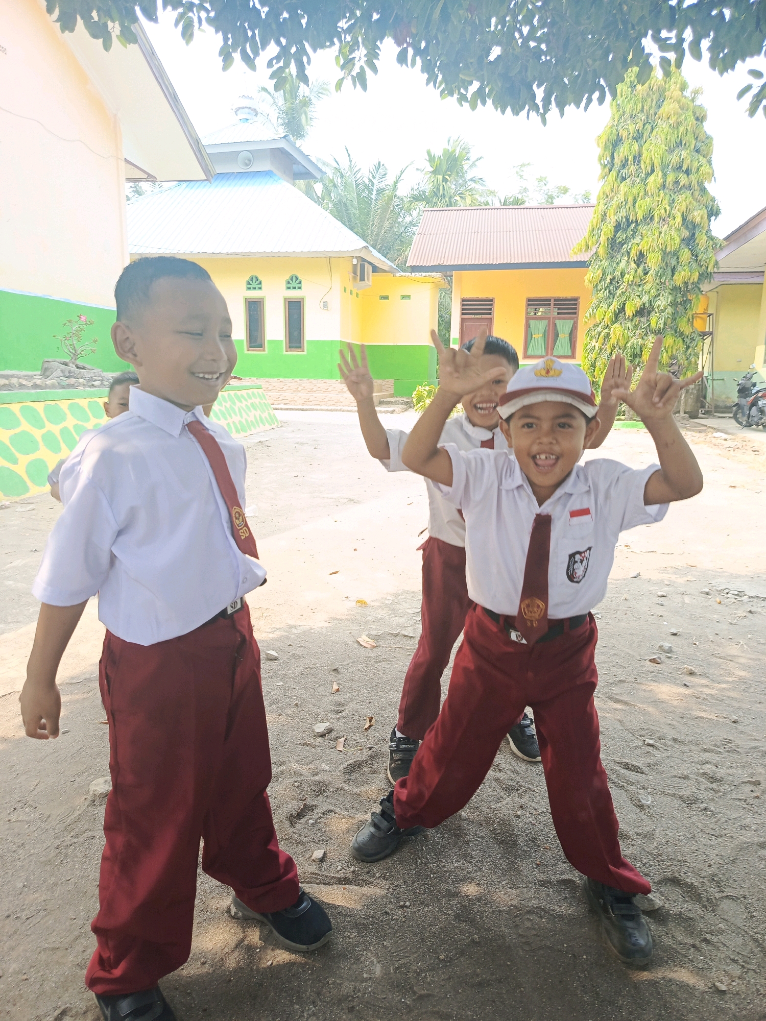 Two young boys, likely elementary school students in Indonesia, are posing for a photo outdoors in what appears to be a schoolyard. The boy in the foreground is wearing a white short-sleeved shirt, a maroon tie with an emblem, and maroon pants. He has a white cap with a red band and emblem. He is smiling broadly and holding both hands up in a gesture with his index fingers and pinky fingers extended. Behind him, another boy in a similar uniform is partially visible, also smiling and holding his hands up in a similar gesture. The background shows school buildings with yellow walls and green trim, some trees, and a clear sky, suggesting a warm, sunny day. The ground is a dirt or gravel surface with some fallen leaves. The scene is likely in Karya Dadi, Indonesia, given the context of Indonesian school uniforms.