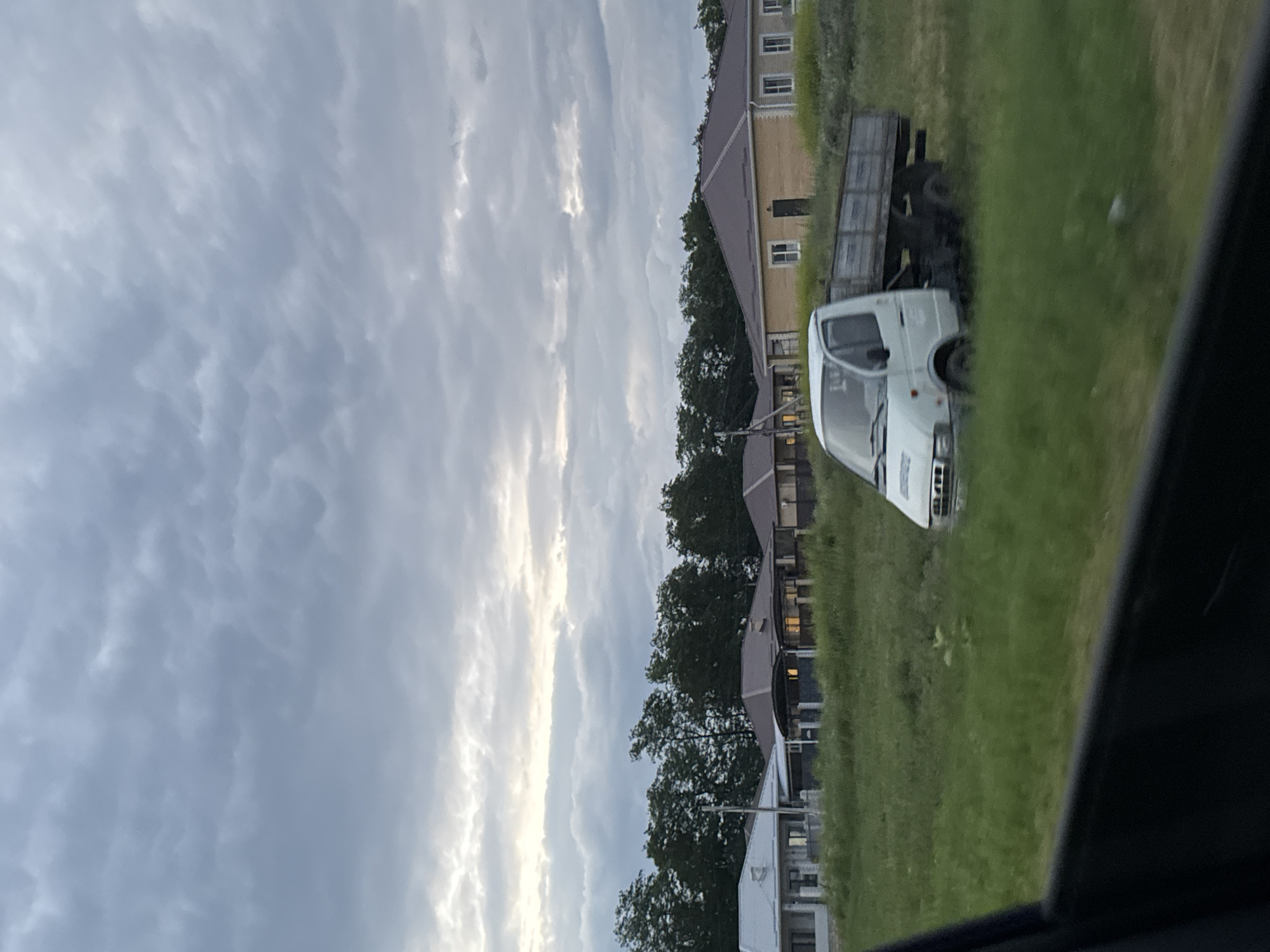 The image depicts a scene with several buildings and a white truck parked on a grassy area. The buildings appear to be modern, with light-colored facades and dark roofs. They are arranged in a row, suggesting a residential or commercial complex. Trees are interspersed between the buildings and along the edge of the scene.
In the foreground, a white truck with a flatbed trailer is visible, parked on the grass. The truck's cabin is white, and it has a dark-tinted windshield. The trailer is dark-colored and appears to be empty.
The sky is overcast with dramatic, textured clouds, suggesting either an impending storm or the aftermath of one. The lighting is soft and diffused, indicating it is likely dusk or dawn. The overall impression is one of a quiet, possibly rural or suburban setting in Mikhaylovsk, Russia. There is no visible text in the image. The scene evokes a sense of calm and perhaps a slight melancholy due to the overcast weather.