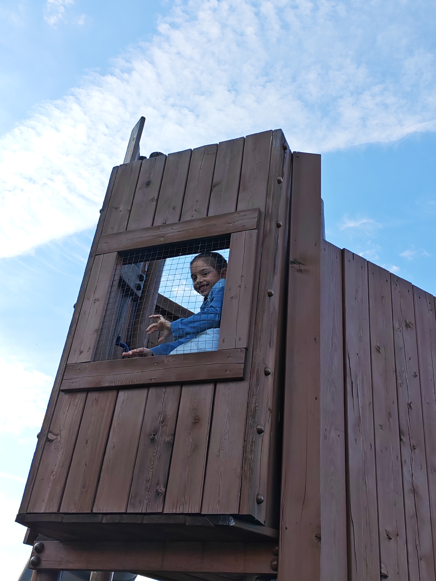 A low-angle shot captures a young person standing inside a wooden structure, possibly a playhouse or lookout tower, set against a blue sky with wispy clouds. The wooden structure is made of vertical planks and has a window with a wire mesh. The young person is visible through the window, smiling and gesturing with their hands. They wear a light-colored shirt and a blue jacket. The location is Bronnitsy, Russia.