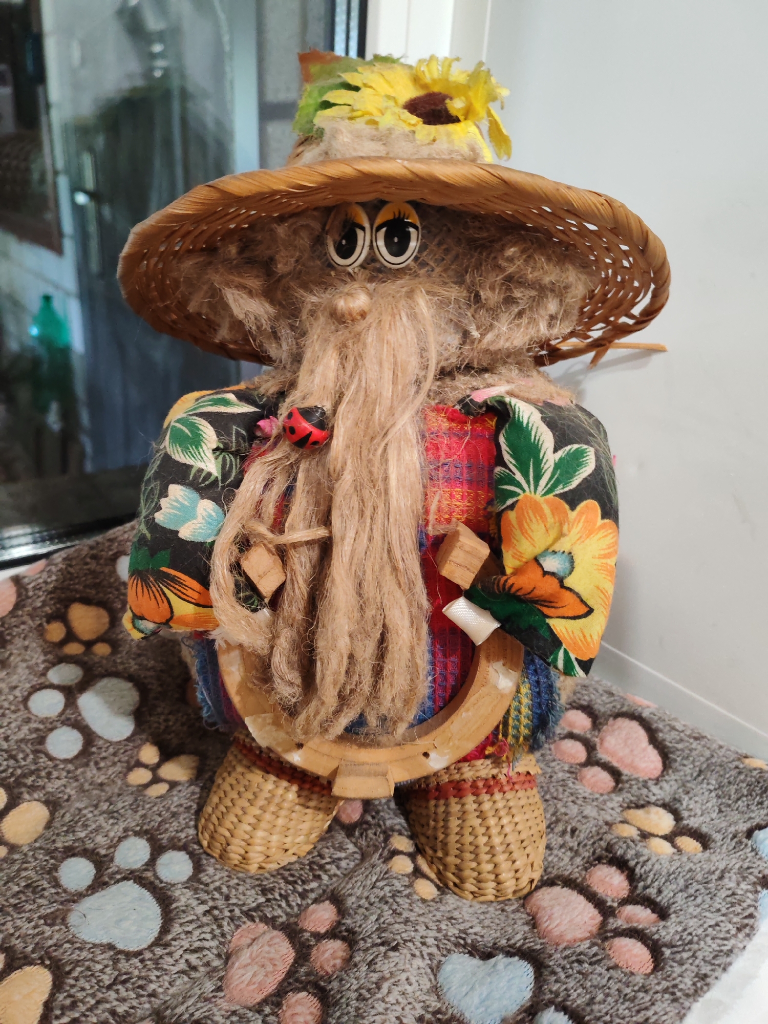 A stuffed doll is pictured standing upright on a grey, plush surface covered in paw prints of various colors. The doll is approximately waist-high and has a large, woven straw hat adorned with a yellow sunflower and green leaves. The doll's face features large, cartoonish eyes with black pupils and white sclera, a small, rounded nose, and a long, stringy, light brown beard.
The doll is wearing a brightly colored, patterned shirt with a black background and floral designs of orange, yellow, and green. Over this, it appears to have a plaid vest or shirt. The doll's lower body is covered in woven straw material, forming legs that end in simple, rounded feet.
Around the doll's neck, a red and black ladybug bead is visible, attached to the beard. Two small, square wooden pieces are also tied to the beard with twine. A light brown, horseshoe-shaped object, made of wood or a similar material, is positioned in front of the doll's lower torso, secured by twine.
The background consists of a light-colored wall to the right and a reflective surface, possibly a window or glass door, to the left. Through the reflective surface, hints of green and possibly other colors of objects are visible, suggesting an indoor setting. The lighting is moderate, with some shadows present. There are no visible people or overt actions taking place; the scene is static and focuses solely on the doll. The location is identified as Тешевичи, Russia.