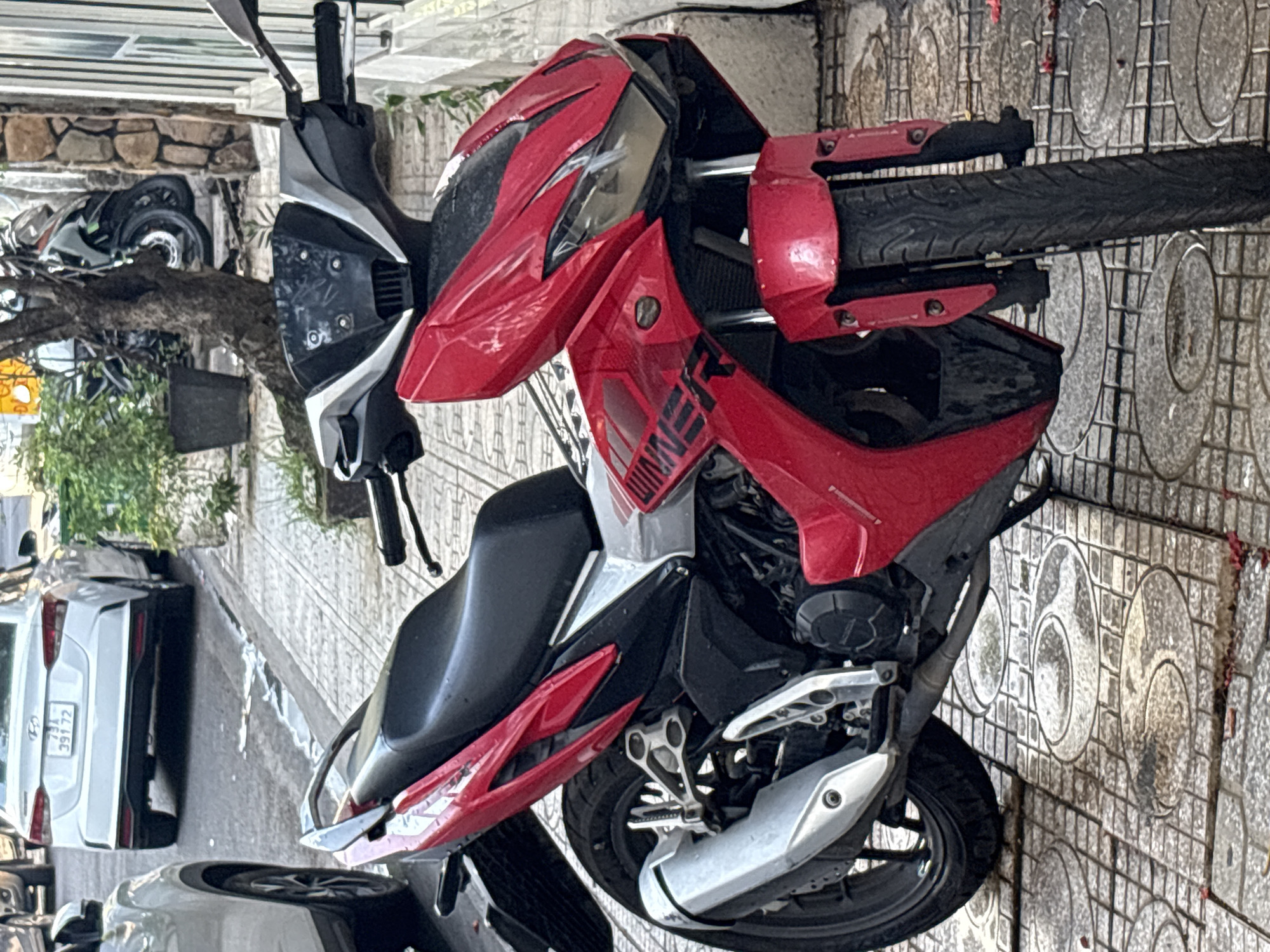 A red and black Honda RS150R motorcycle is parked on a tiled sidewalk in front of a building. The motorcycle has a sporty design with sharp lines and a silver exhaust pipe. The sidewalk is made of square tiles with circular patterns. In the background, a white car is parked on the street. The scene appears to be in a city in Vietnam, as suggested by the license plate on the white car. The weather appears to be overcast, with some puddles of water on the street. The image is taken from a slightly low angle, looking up at the motorcycle.